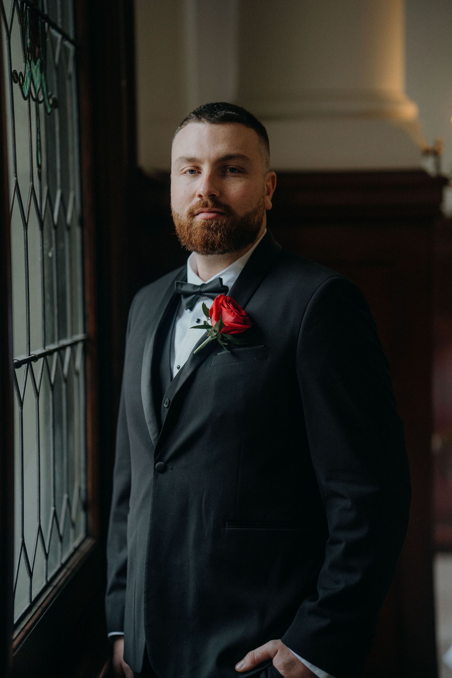 Groom in black tuxedo with red rose boutonniere stands by window in elegant venue.