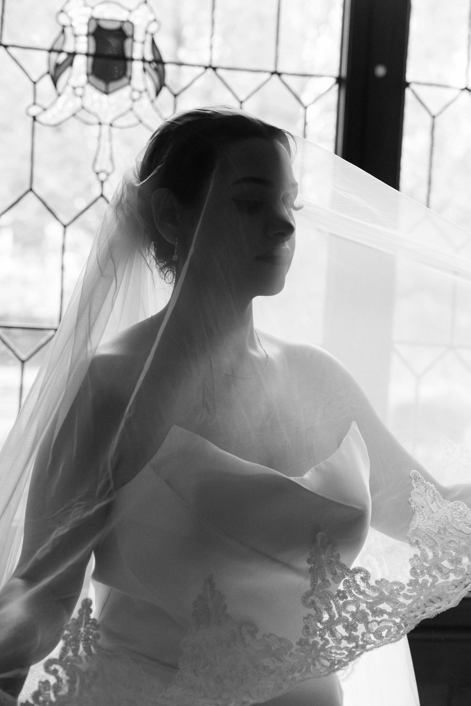 Black and white portrait of a bride in a strapless lace gown and flowing veil, standing before a stained glass window.