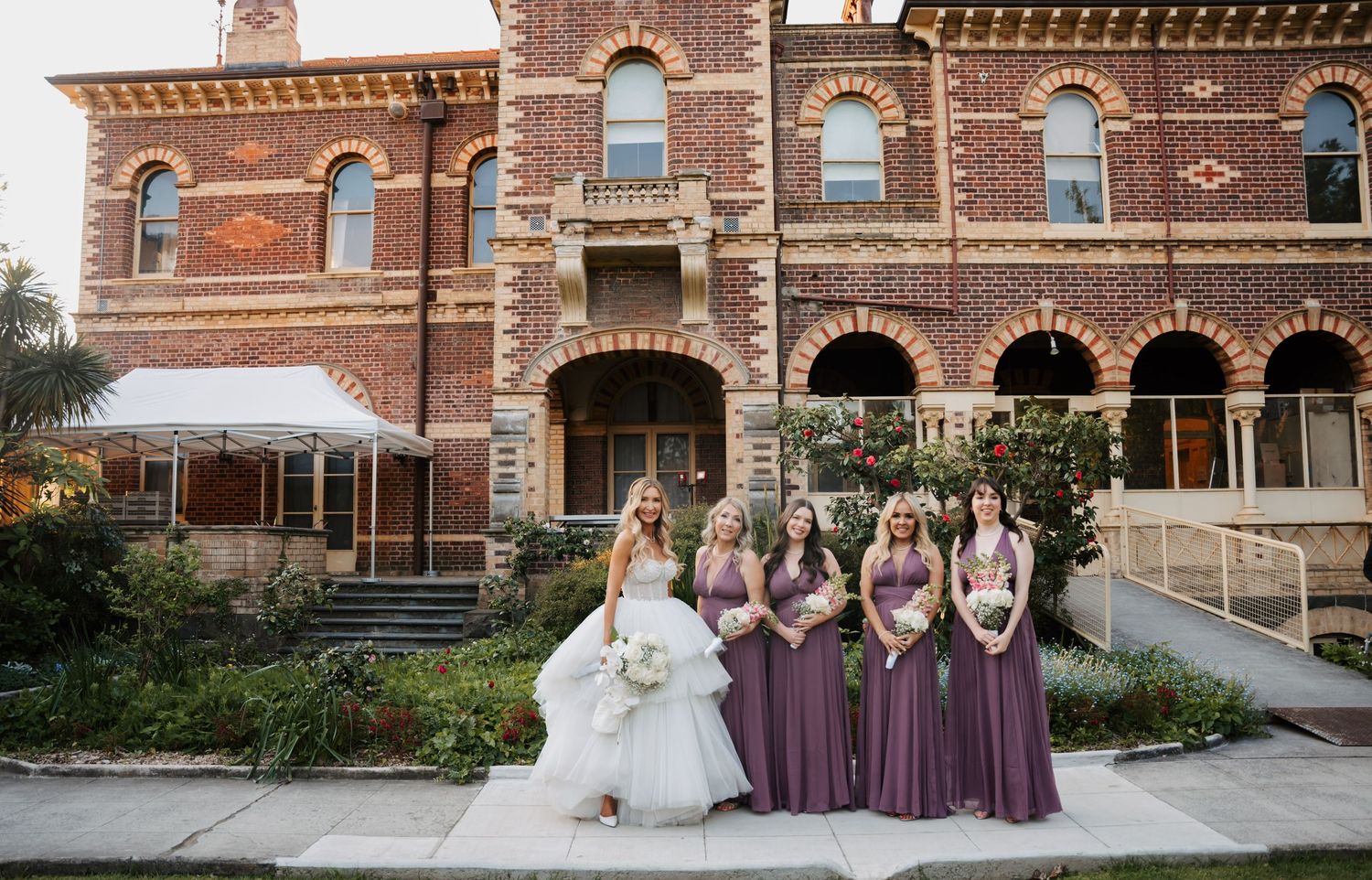 Wedding party poses in front of historic brick building with ornate Victorian architecture and landscaped grounds.