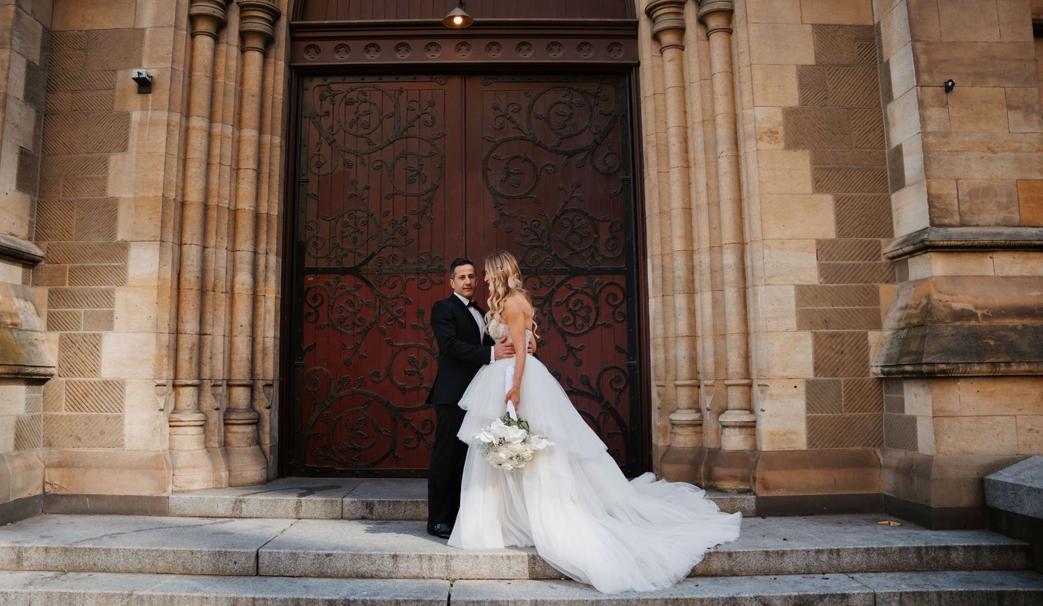 A couple in wedding attire stands together on stone steps in front of an ornate wooden church door.
