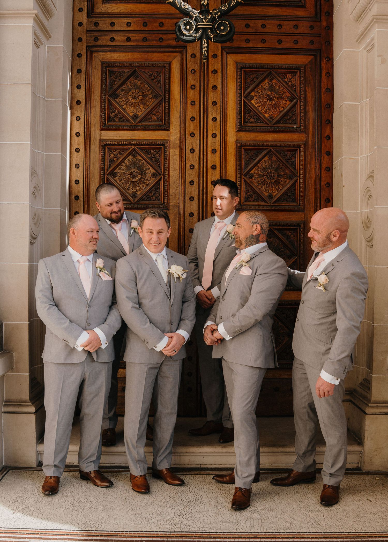 Wedding party in light gray suits poses playfully in sequence against ornate wooden doors at elegant venue.