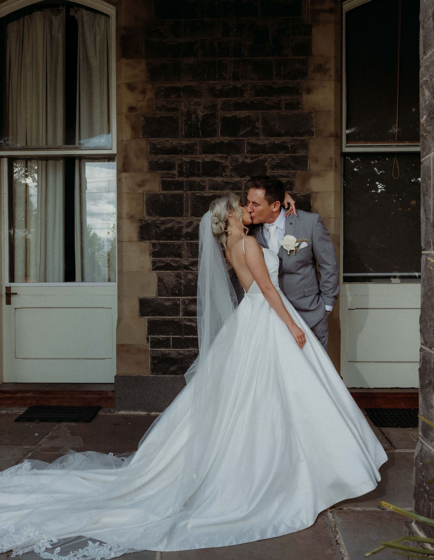 A romantic wedding couple shares an intimate moment on a stone porch with a flowing white dress train.