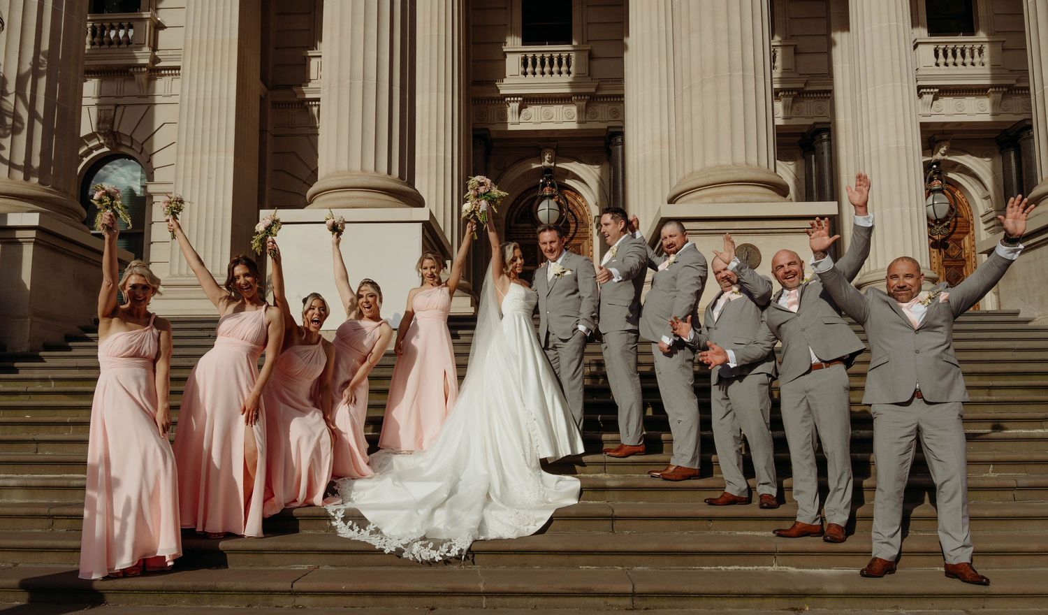 Wedding party members in pink dresses and gray suits pose on grand stone steps of a historic building.