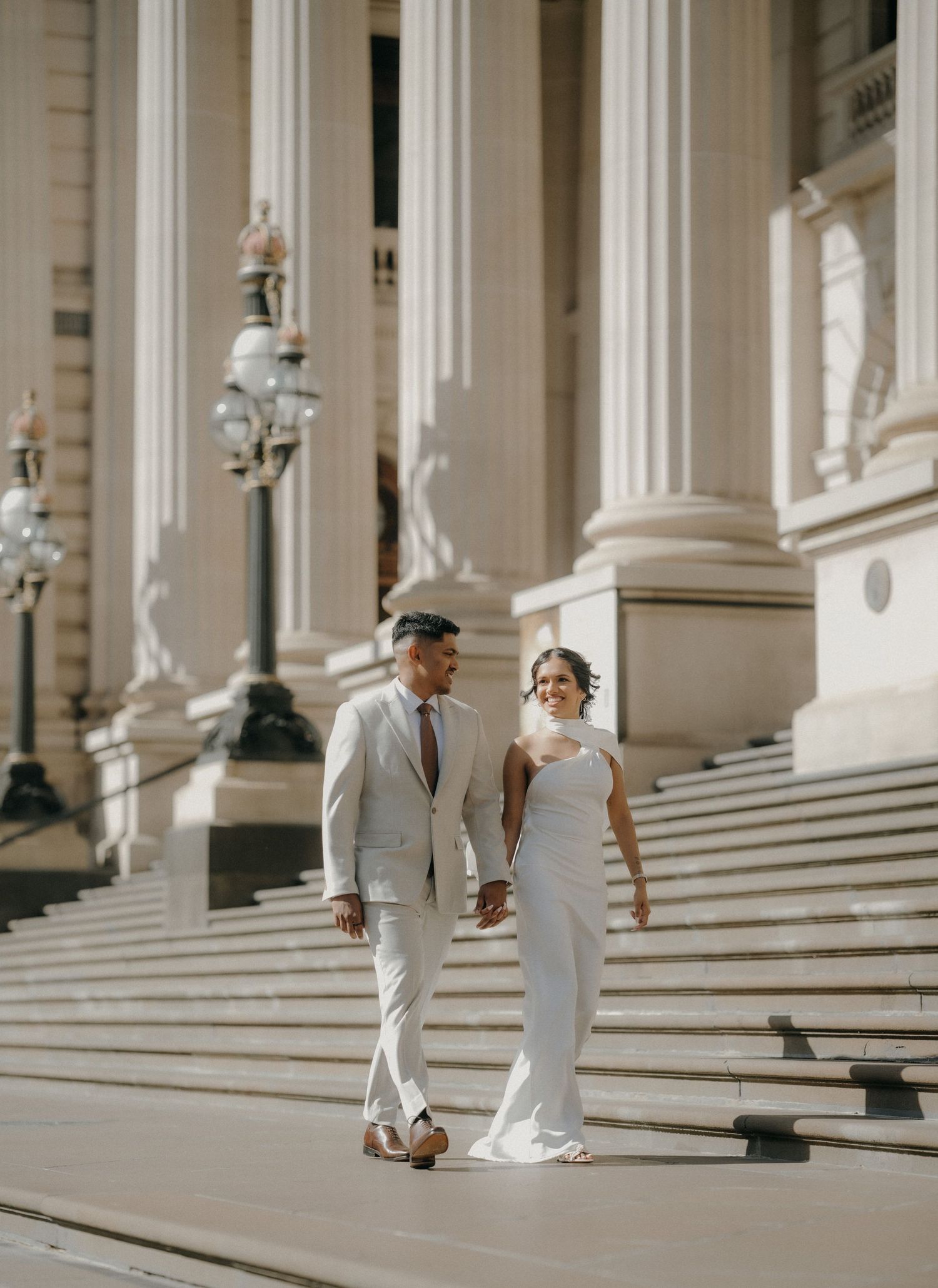 A couple in elegant white attire walks gracefully up grand marble steps of a classic architectural building.