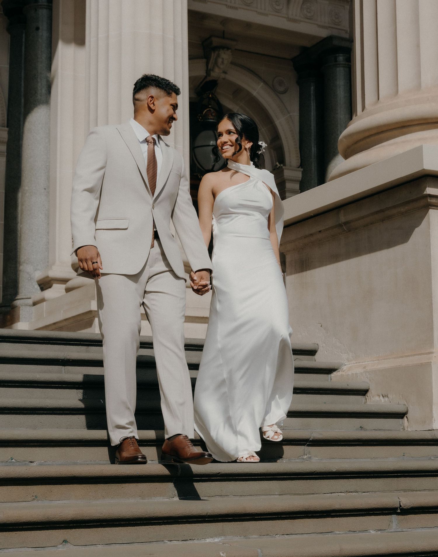 A couple in white wedding attire walking up elegant stone steps together at a grand venue.