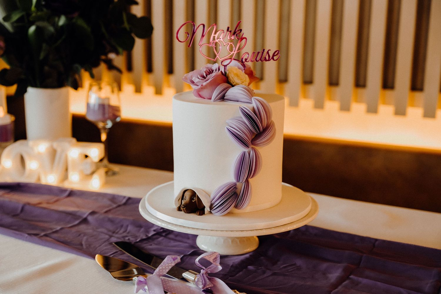 A wedding cake decorated with cascading purple ribbon and delicate floral accents sits elegantly on a white stand.