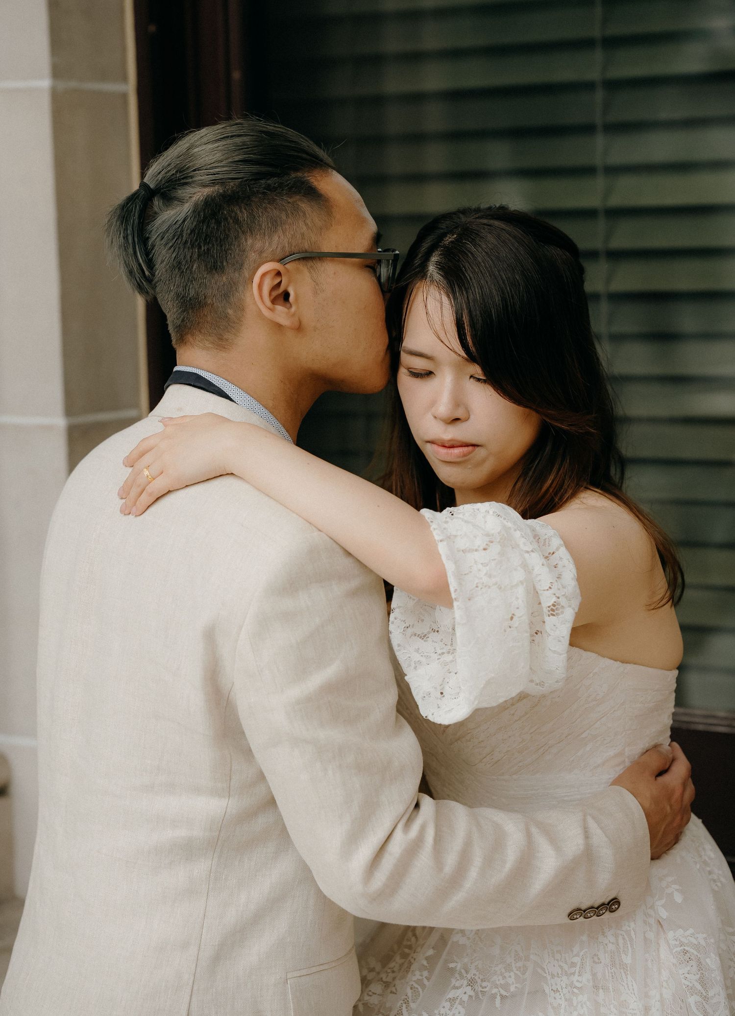 A tender romantic moment between newlyweds in white wedding attire embracing near window blinds.