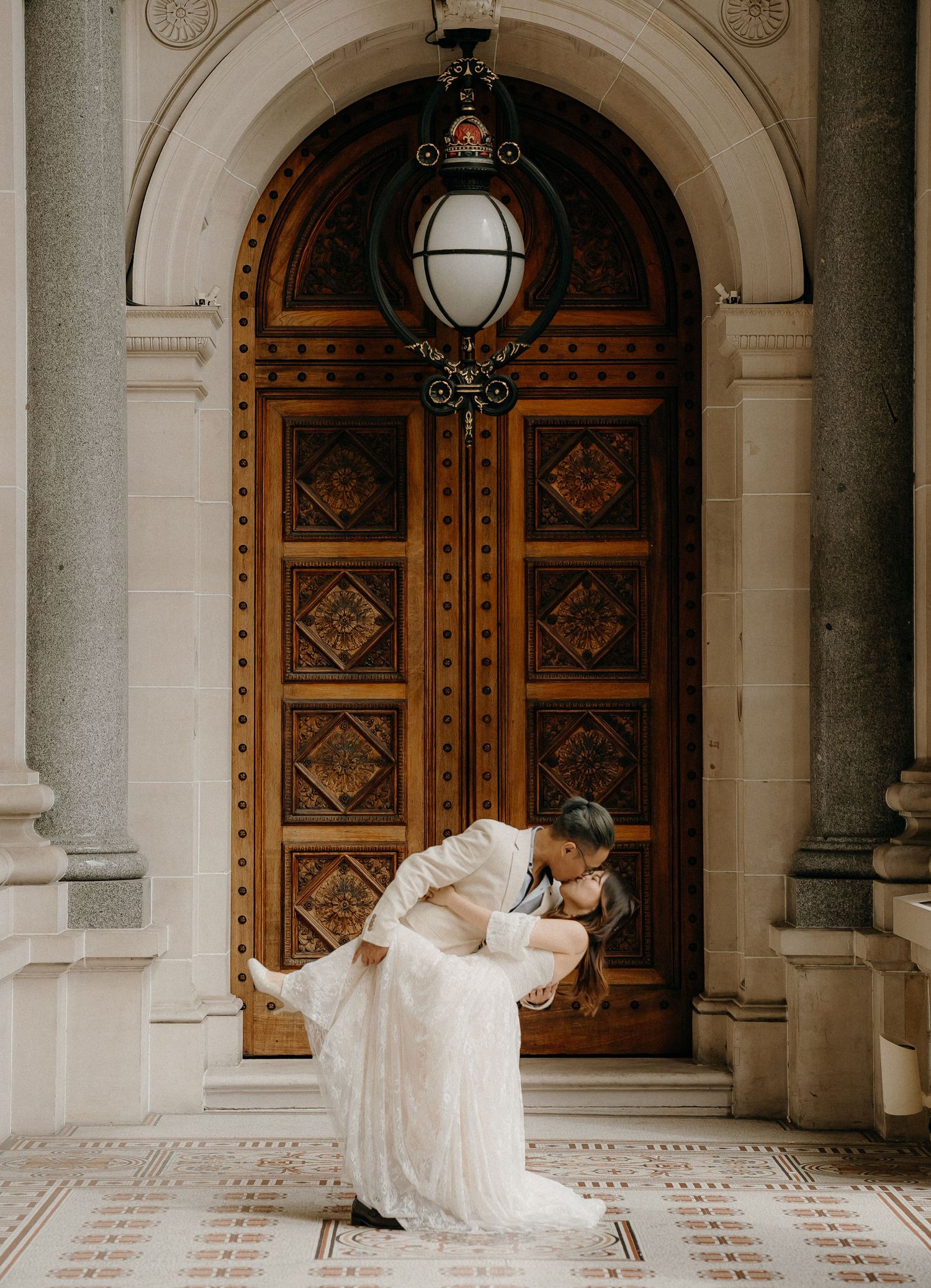 Elegant couple in formal attire share romantic dip pose in front of ornate wooden door with vintage hanging lamp.