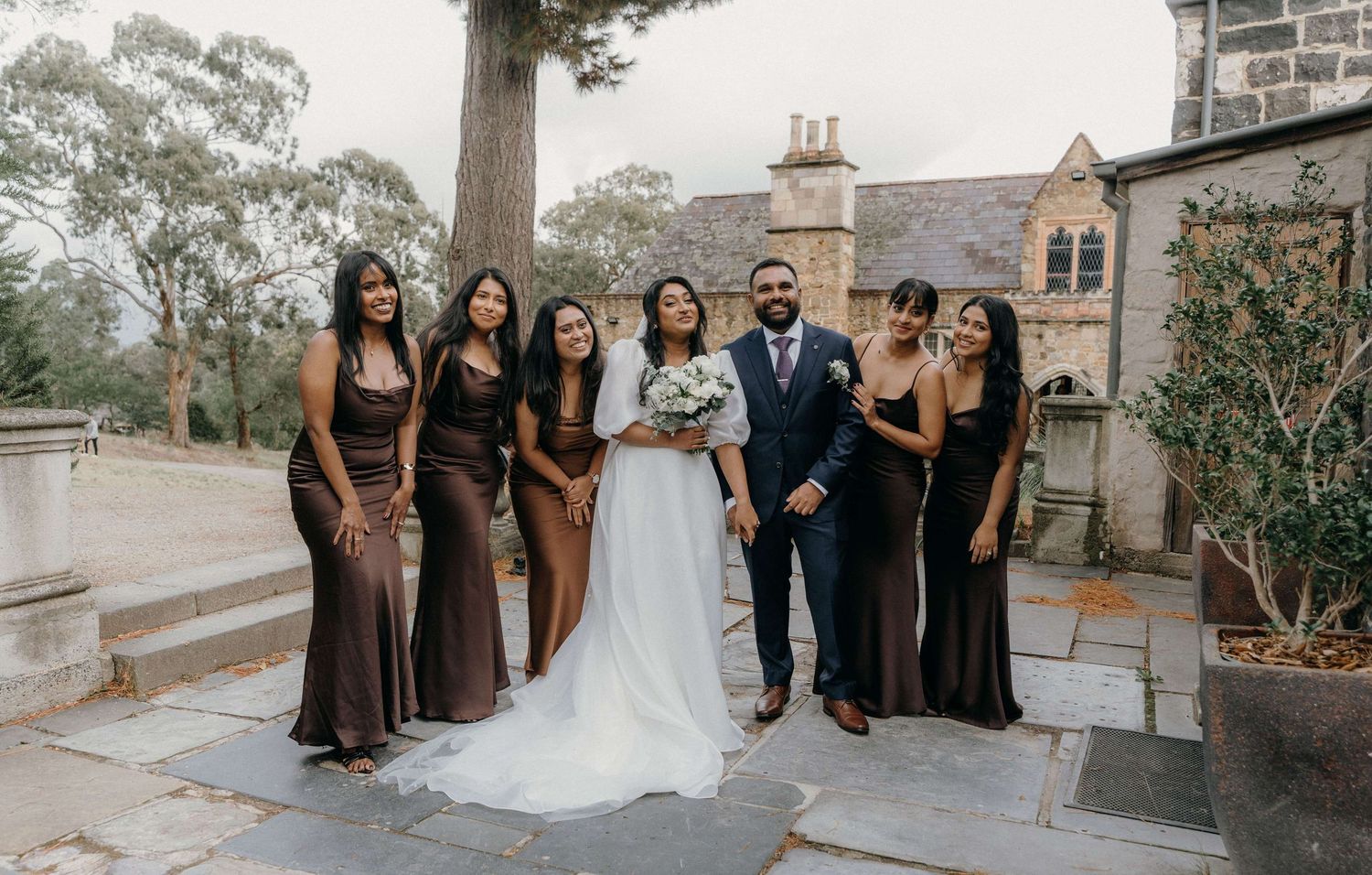 Wedding party poses together on stone patio outside historic brick church building in elegant formal attire.