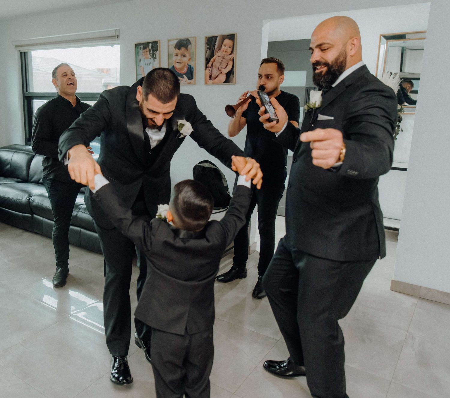 A group in formal black suits dance and celebrate together in a bright room with photographs on the wall.
