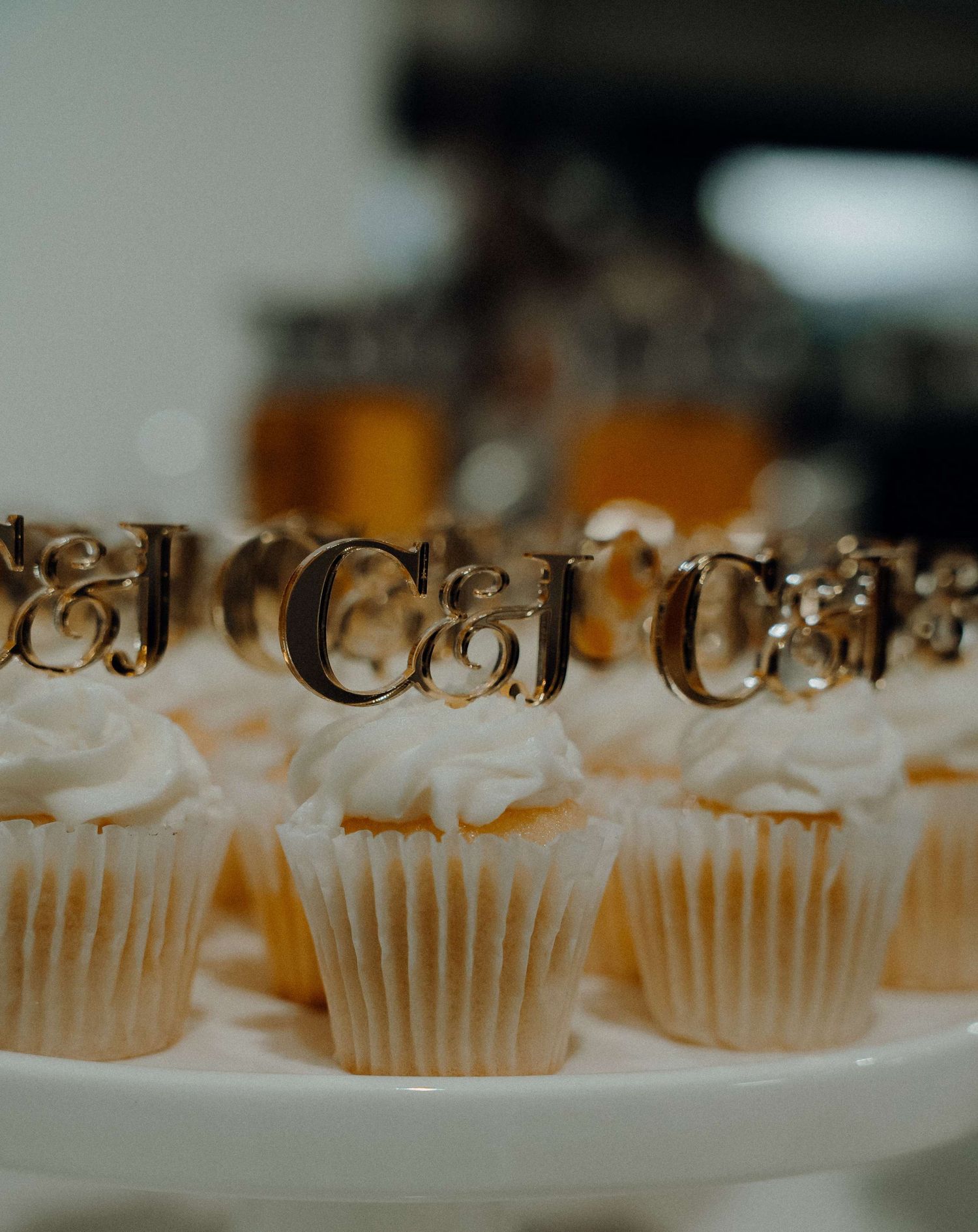 Row of white frosted cupcakes decorated with gold script letters arranged on a table with shallow depth of field.