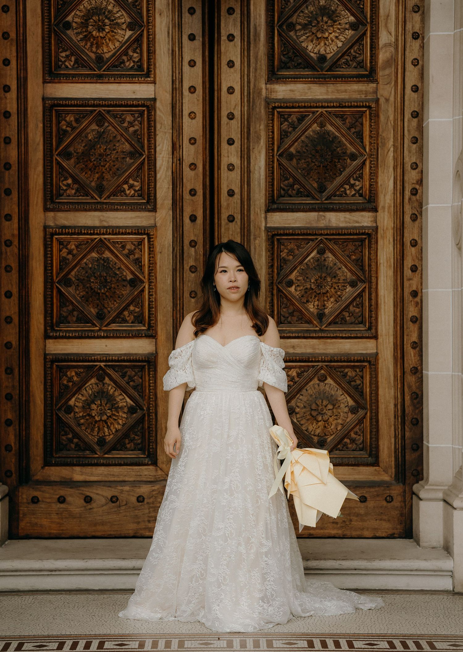 Elegant bride in a flowing white lace dress poses against ornate wooden doors with decorative panels.