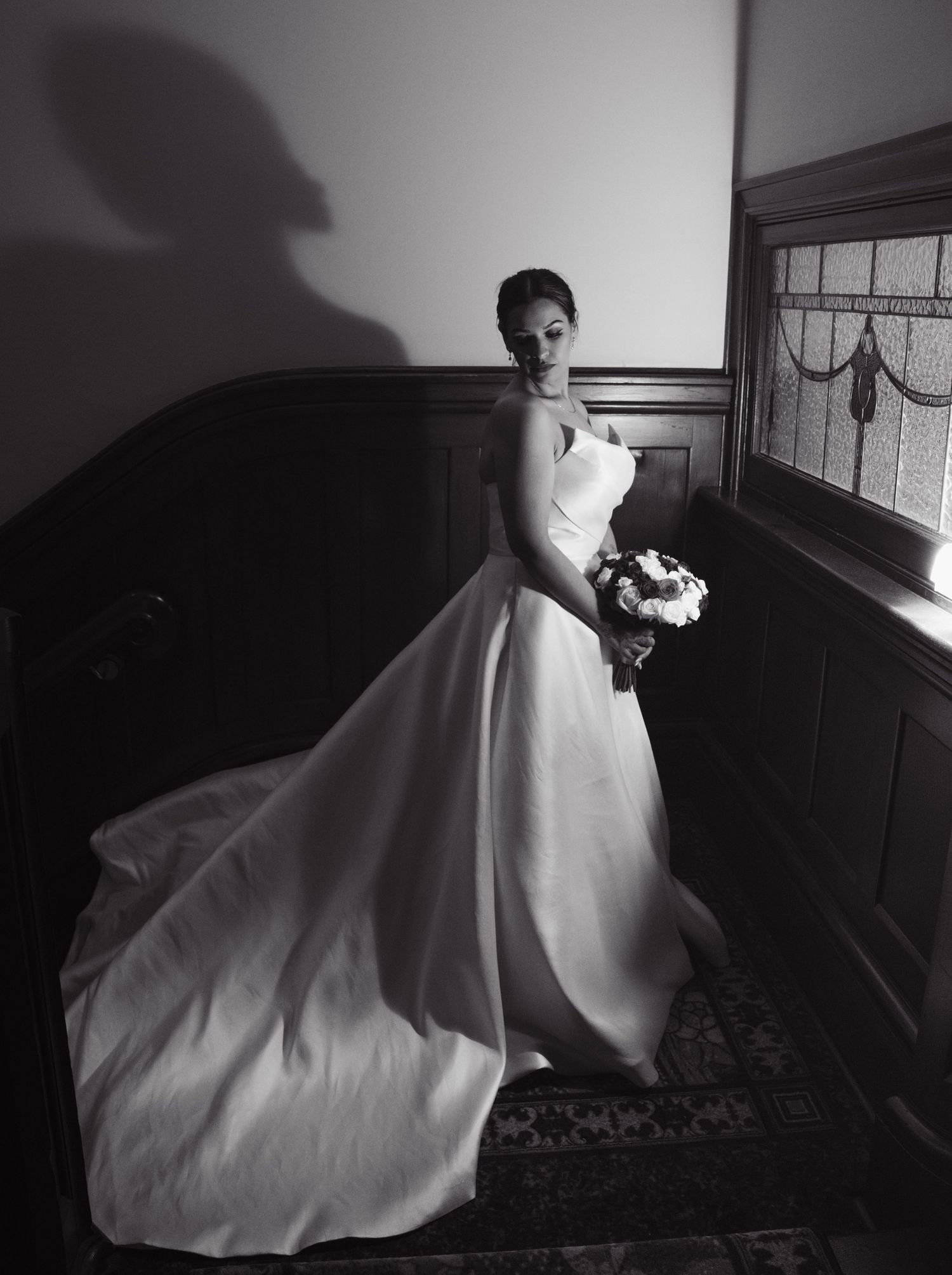 Bride in elegant white ball gown with long train holds bouquet on staircase in black and white wedding photo.