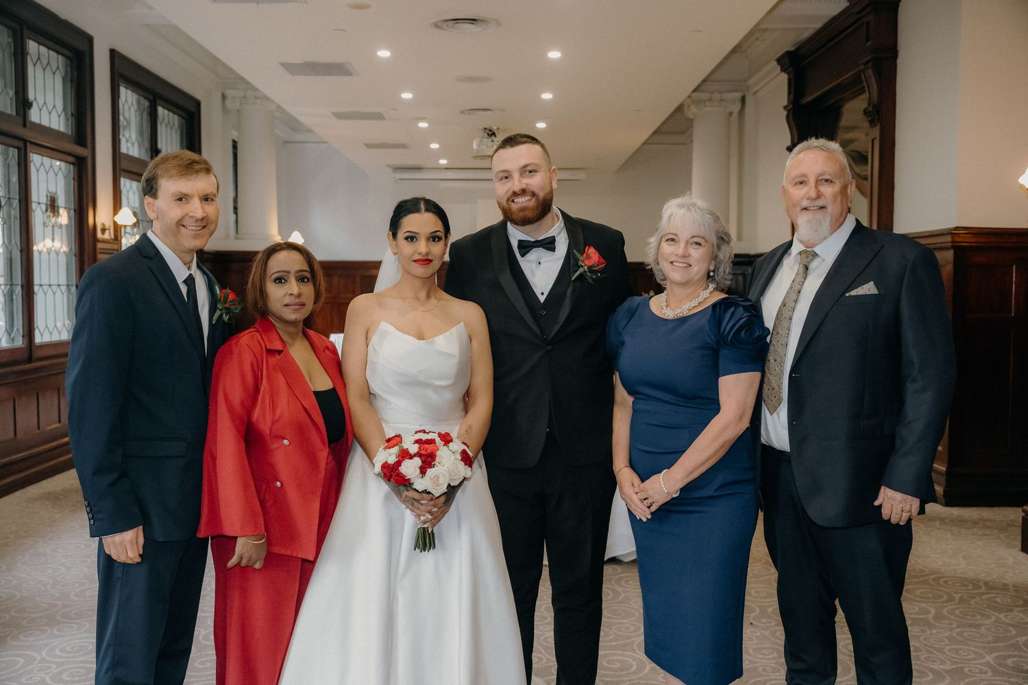 Wedding party of six posing indoors, bride in white gown holding red bouquet with groom in black tuxedo and family.