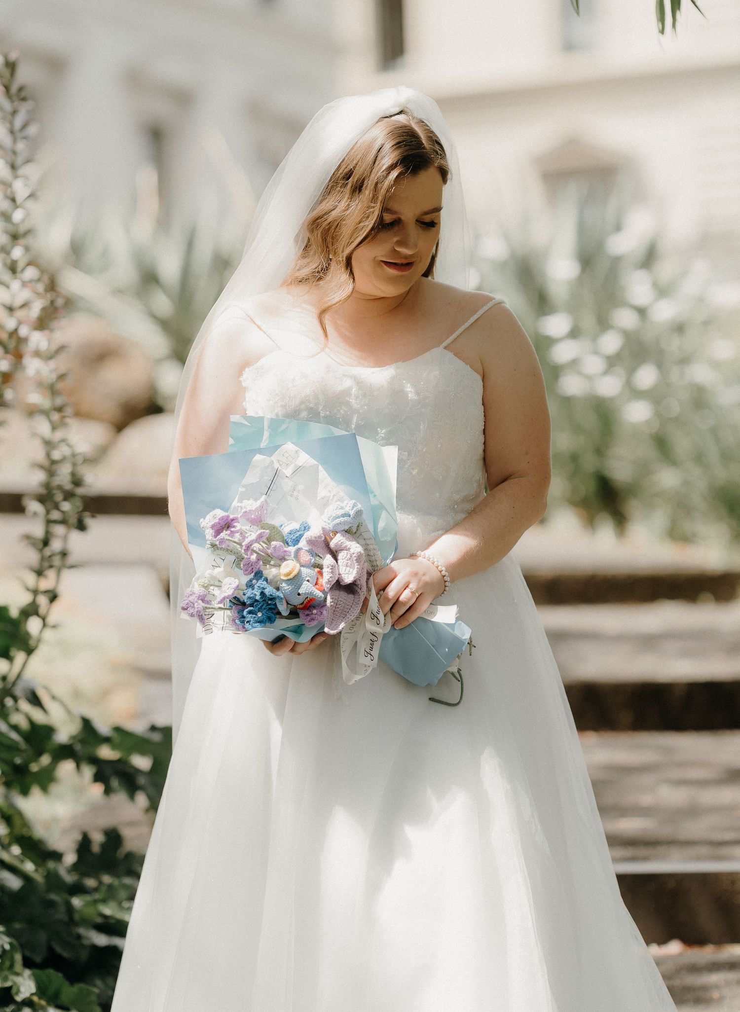 A bride in a flowing white strapless gown holds a blue and purple bouquet while standing in a garden setting.