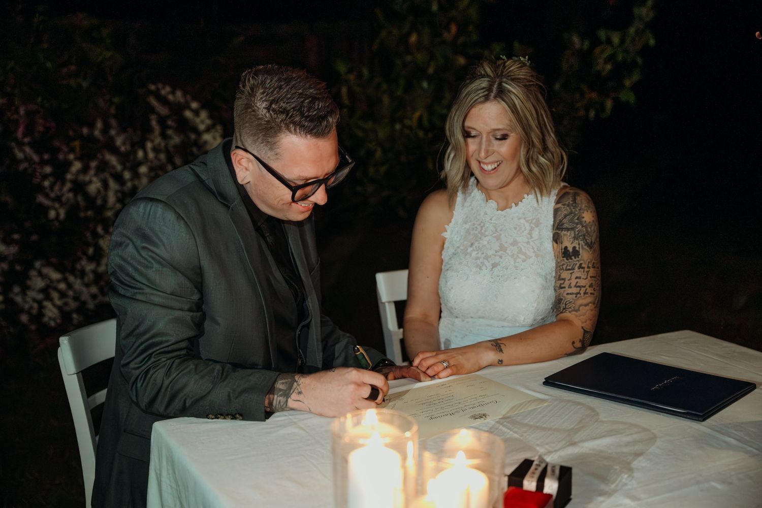 A couple sits at a candlelit table during an evening celebration with paperwork and candles visible.