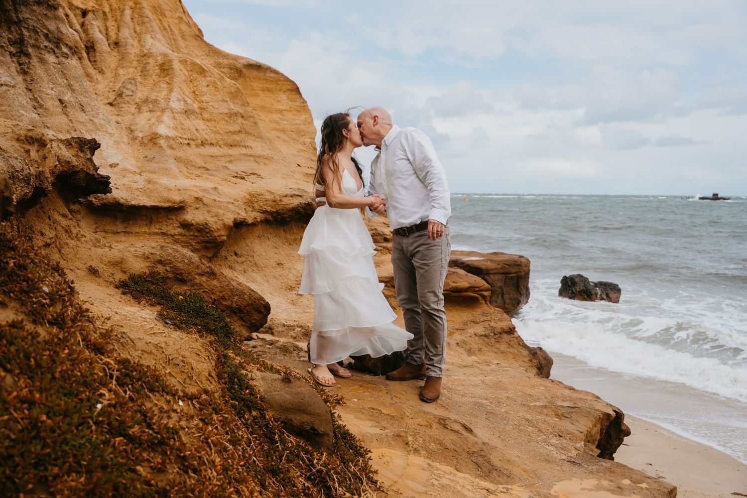 A romantic wedding couple shares a moment on a rocky cliff overlooking a beach at sunset.