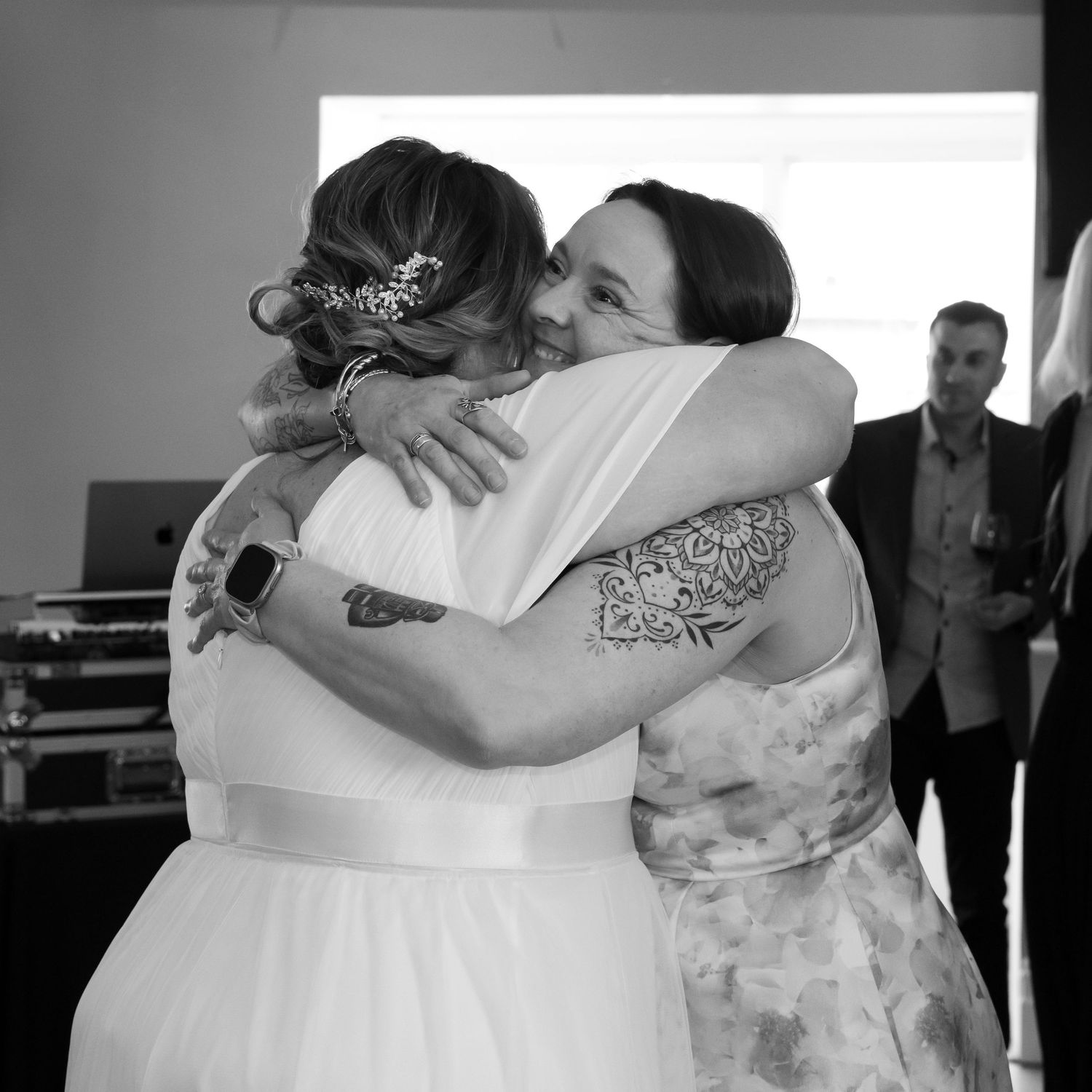 Two people share an emotional embrace at a wedding celebration, with a guest visible in the background.