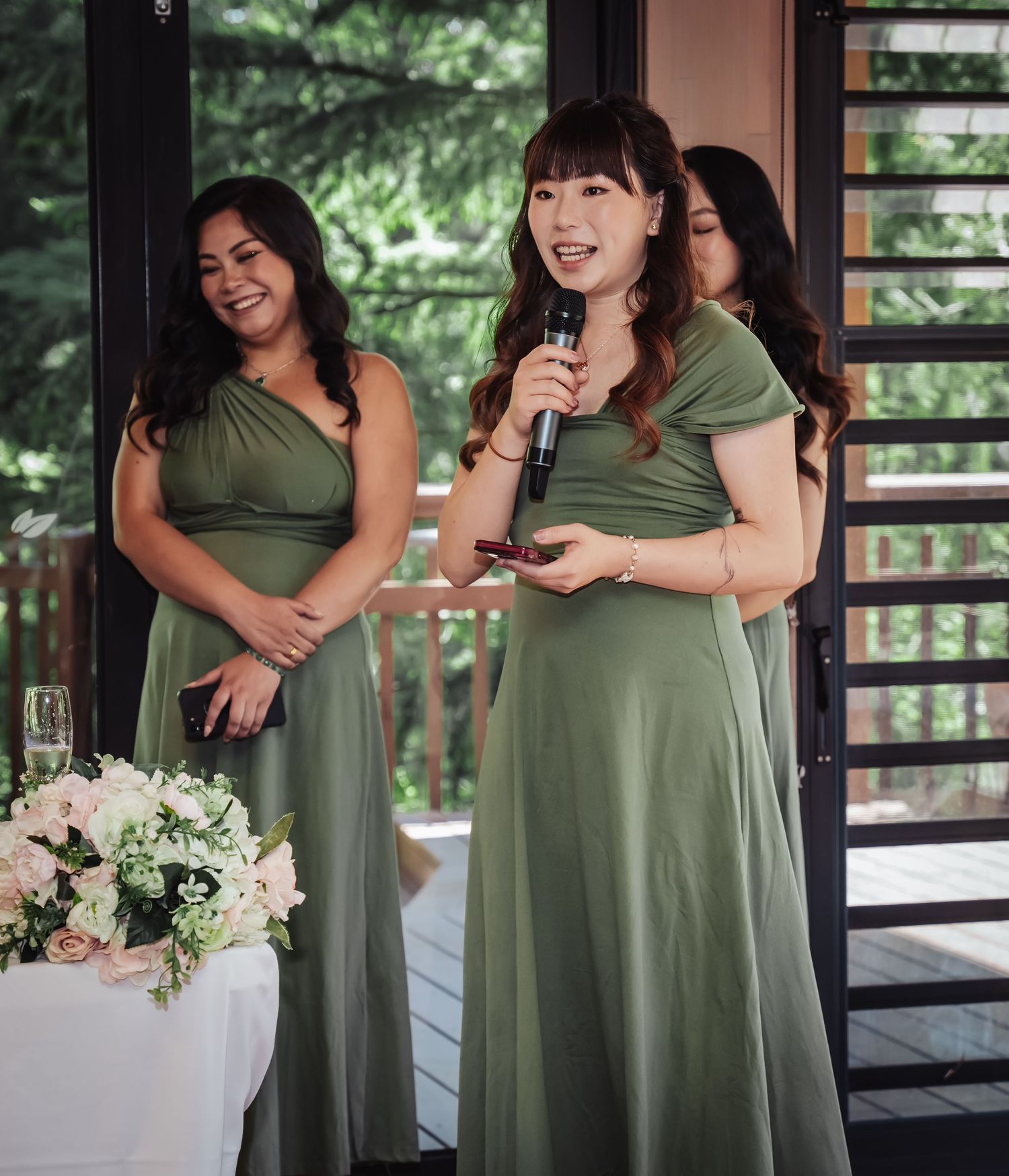 Two bridesmaids in sage green dresses share a candid moment while holding white floral bouquets on a porch.