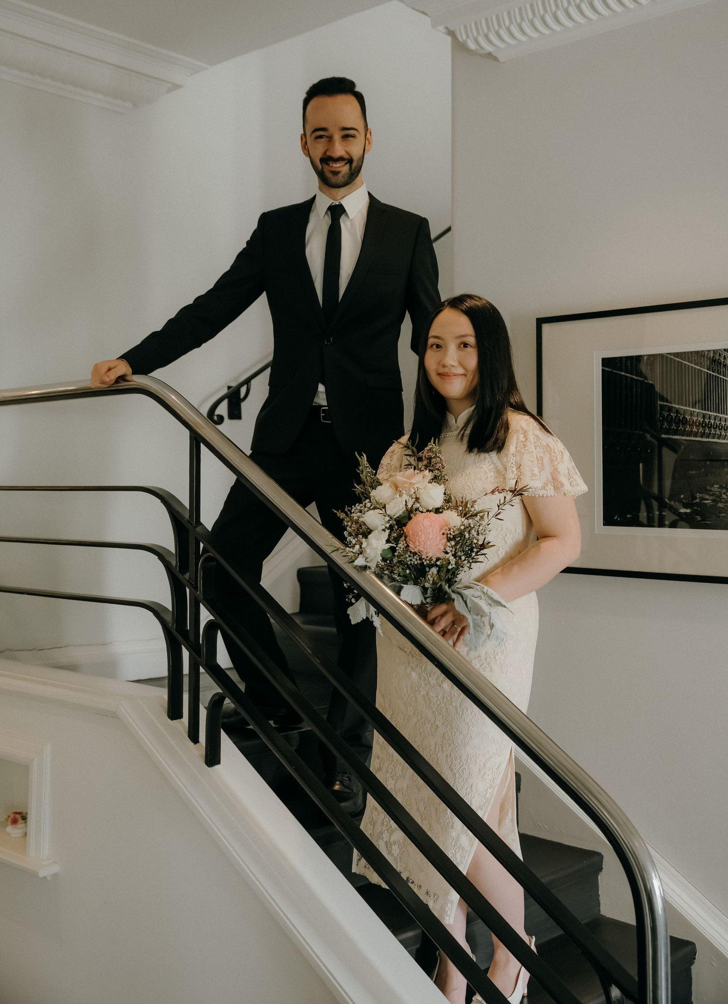 Bride and groom posing on staircase, bride holding pink bouquet in lace dress, groom in black suit.