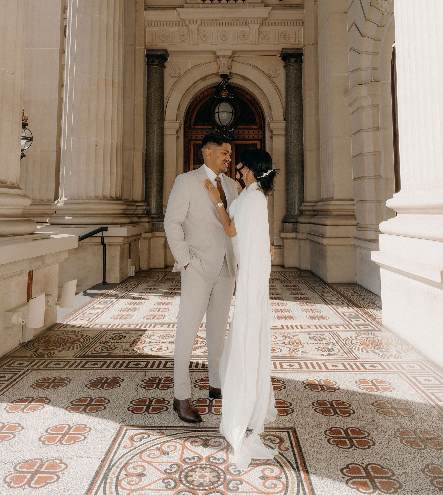 A couple in formal wedding attire embraces in a grand marble hallway with ornate floor patterns and columns.
