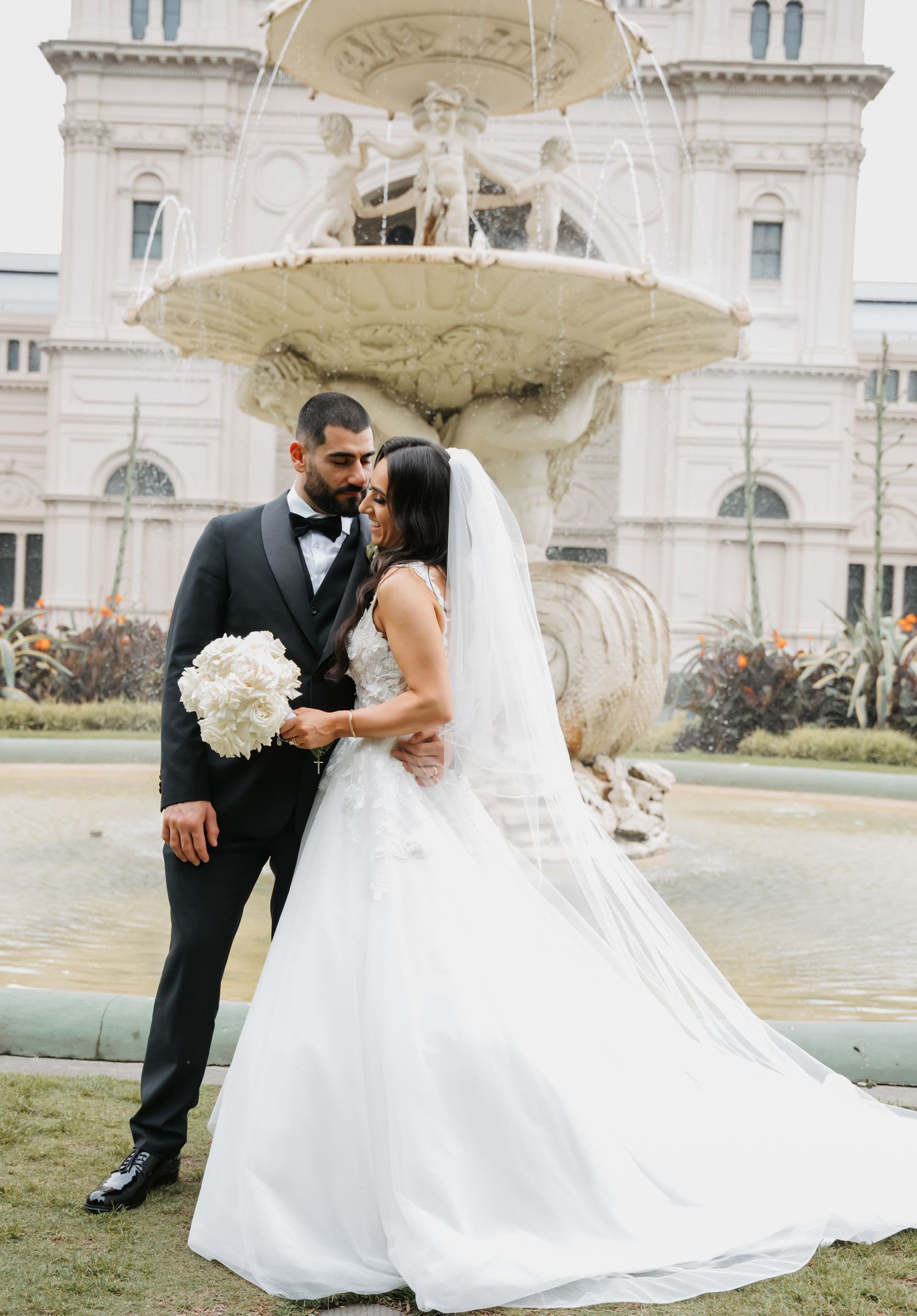 A bride and groom pose by an ornate fountain outside a grand historic building in their wedding attire.
