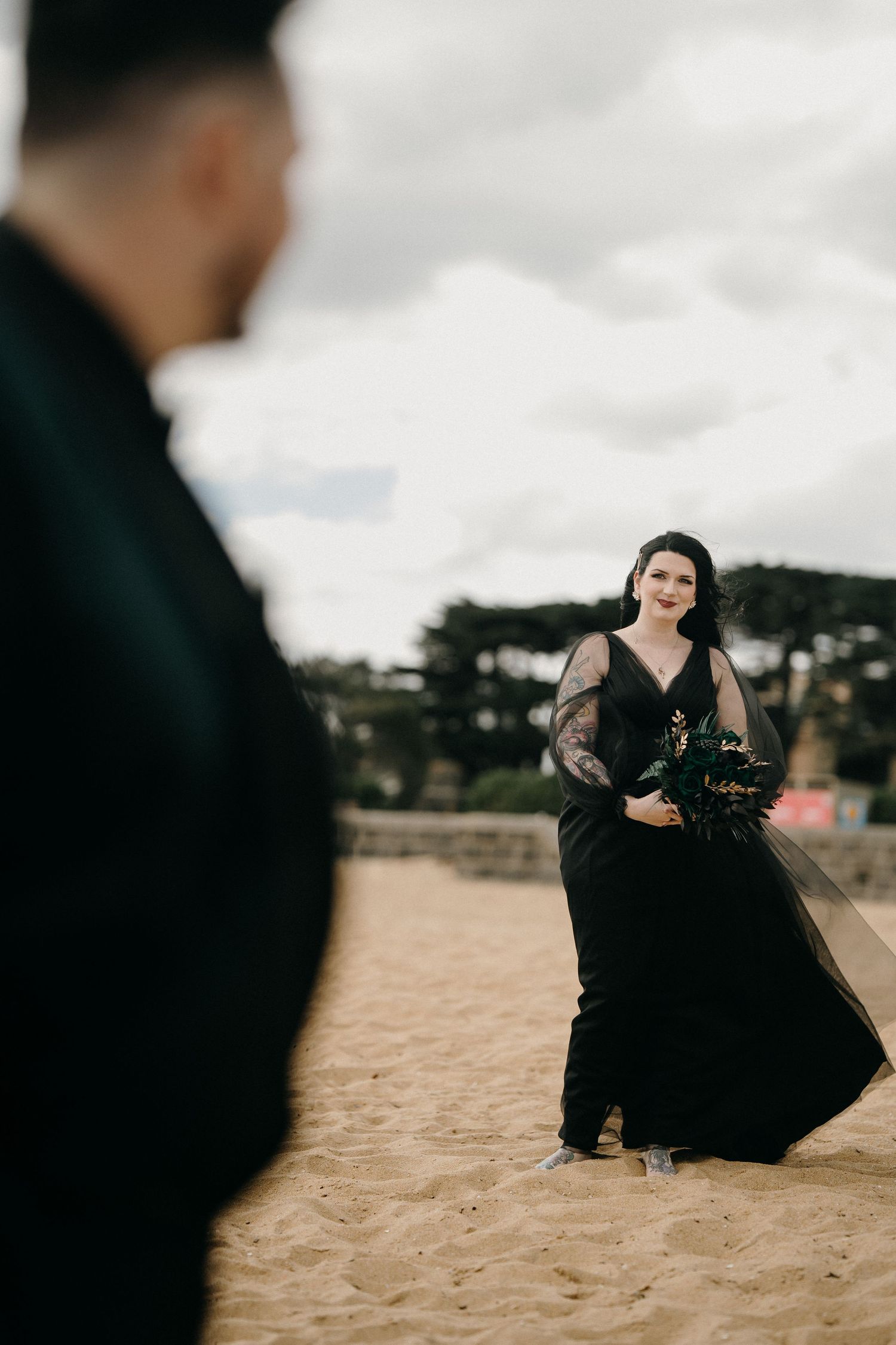 A figure in a flowing black dress poses dramatically on a sandy beach against a cloudy sky.