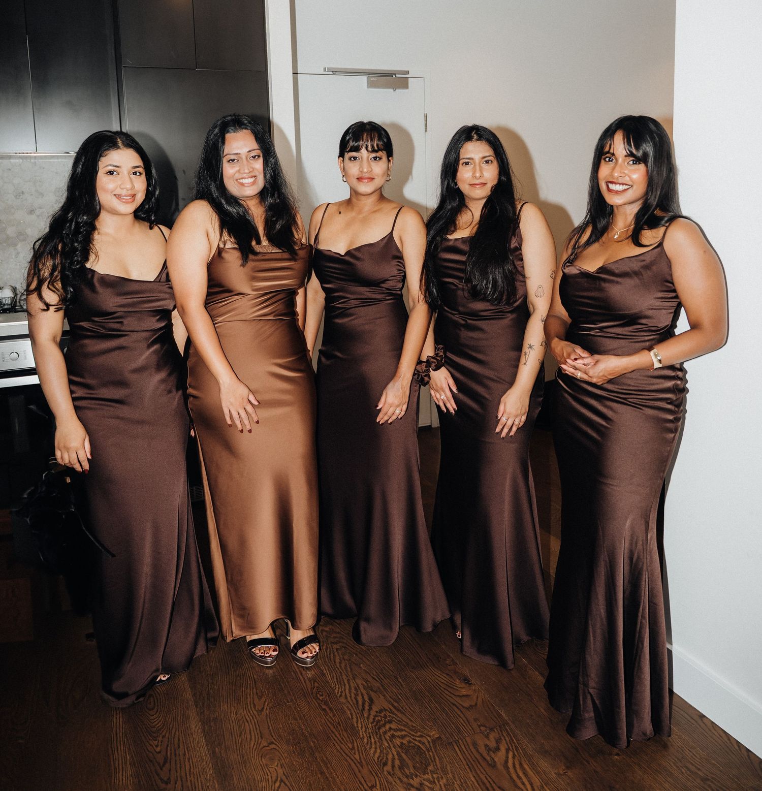 Five bridesmaids pose together wearing elegant chocolate brown satin gowns in a wedding party photo.