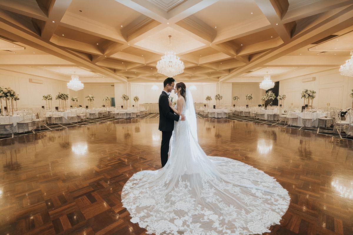 A grand ballroom with wooden floors and elegant chandeliers showcases a flowing white wedding gown with dramatic cathedral train.