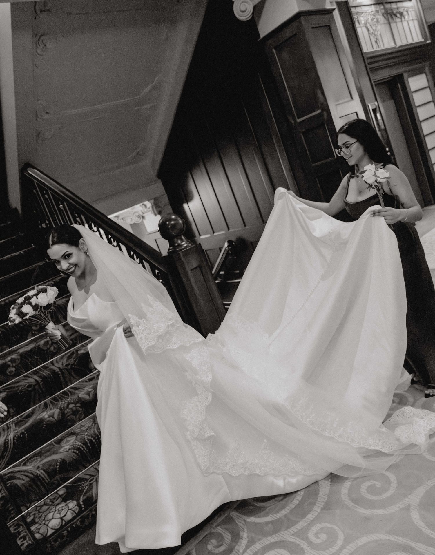 Black and white photo of a bride in a flowing gown on stairs as bridesmaid lifts her long train.
