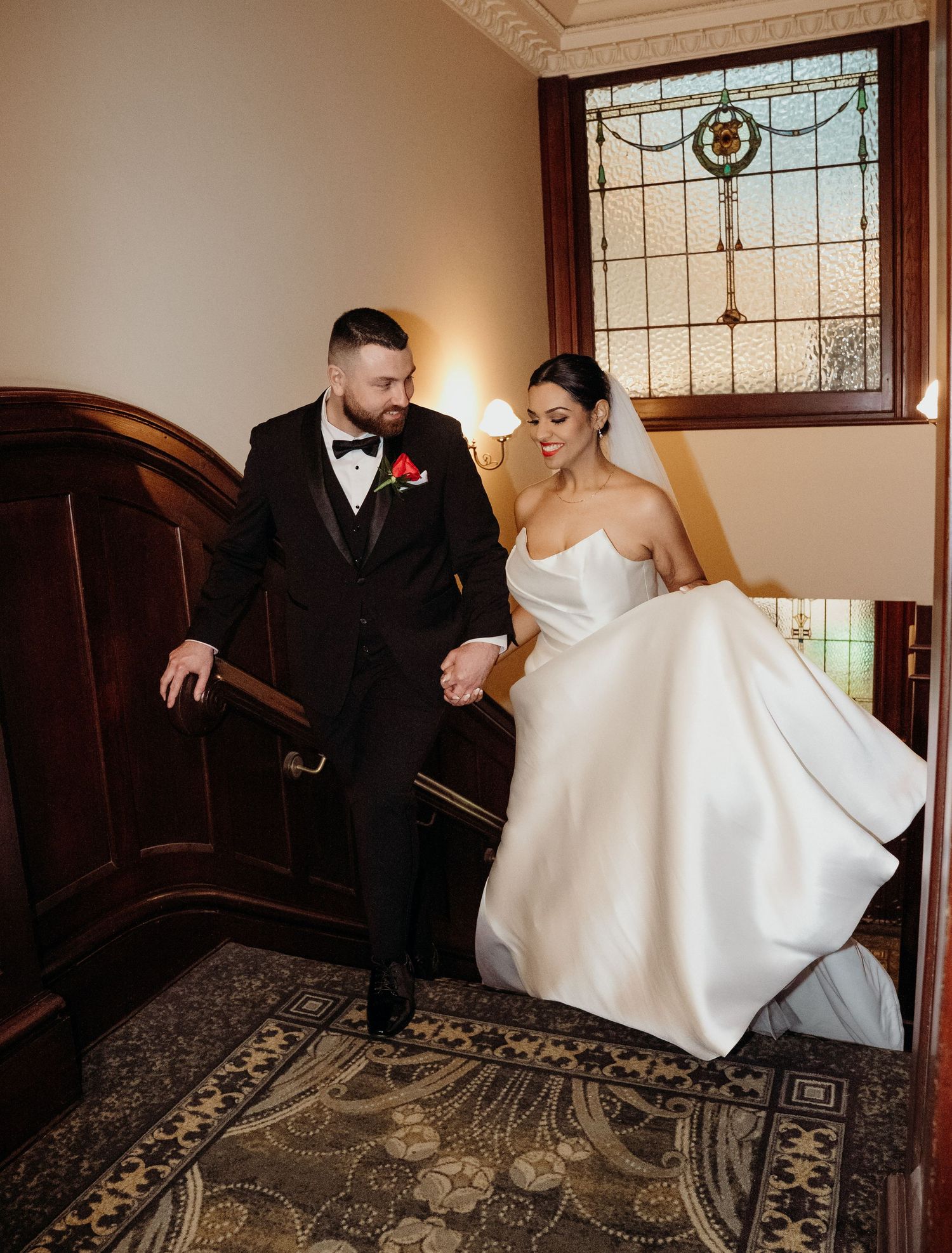 A bride and groom hold hands while walking up an elegant staircase, with stained glass windows in the background.