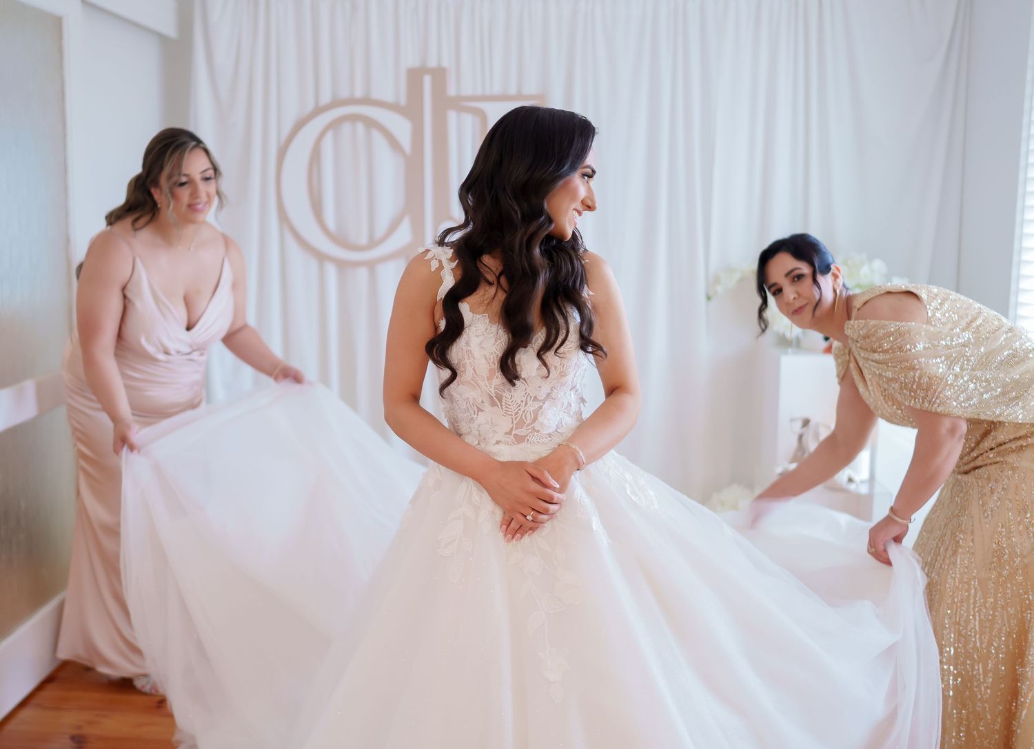 Three people in formal gowns prepare in a bright room with white drapery, with one adjusting a flowing white dress.