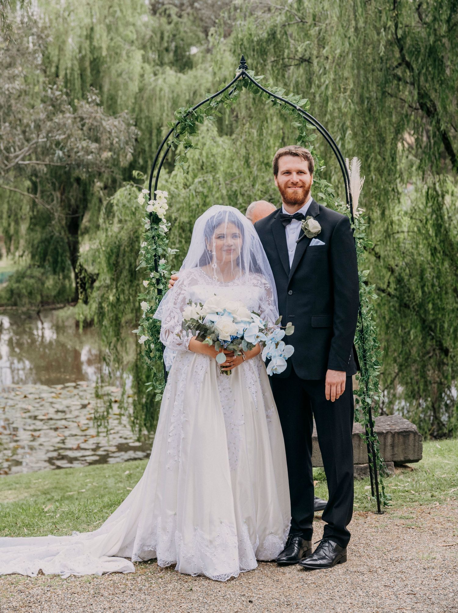 A couple stands under a green arch on their wedding day beside a pond with weeping willow trees in the background.