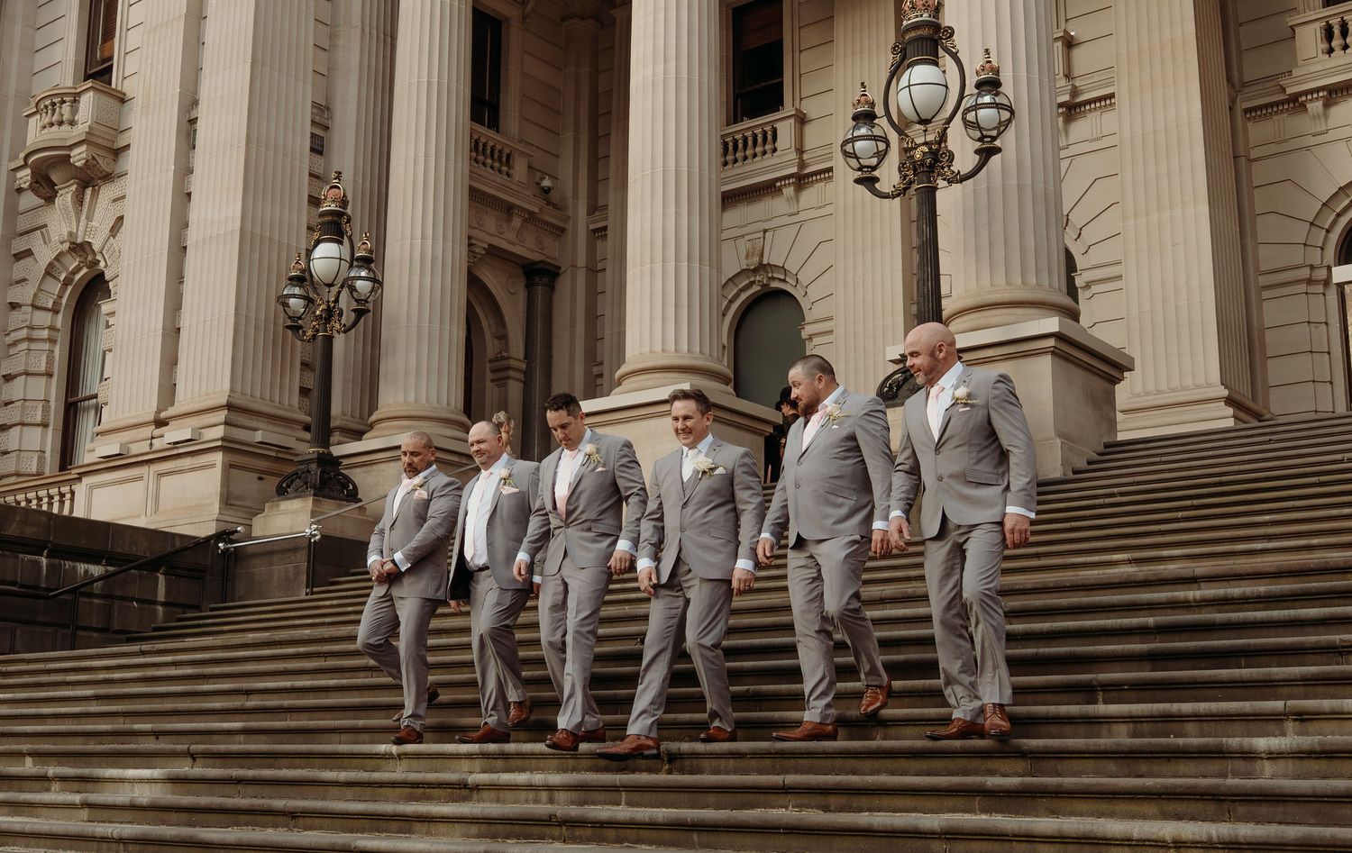 Wedding party in gray suits poses on grand stone steps of historic building with ornate columns and lamp posts.