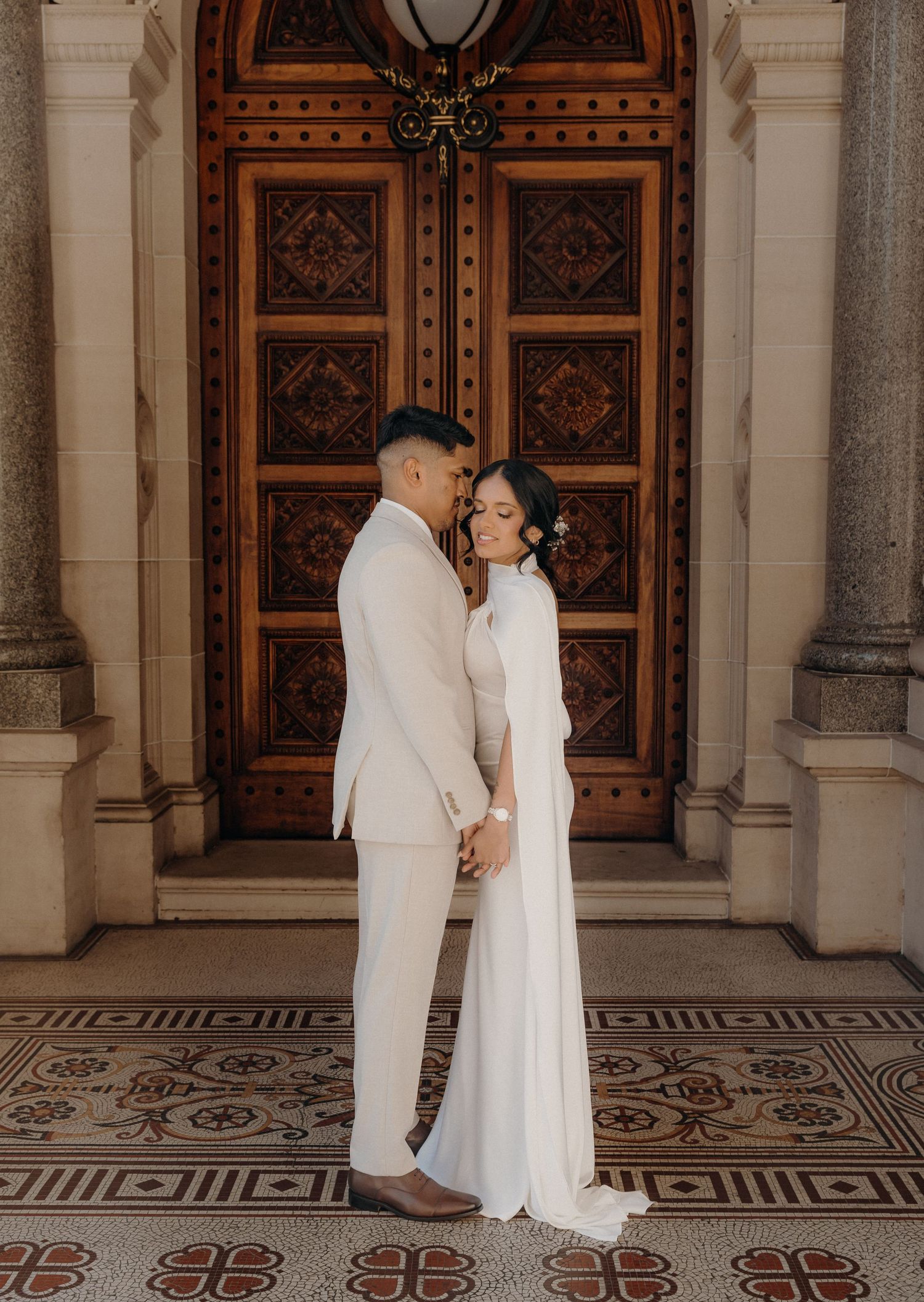 Elegant couple in matching white attire share intimate moments in front of ornate wooden doors at historic venue.
