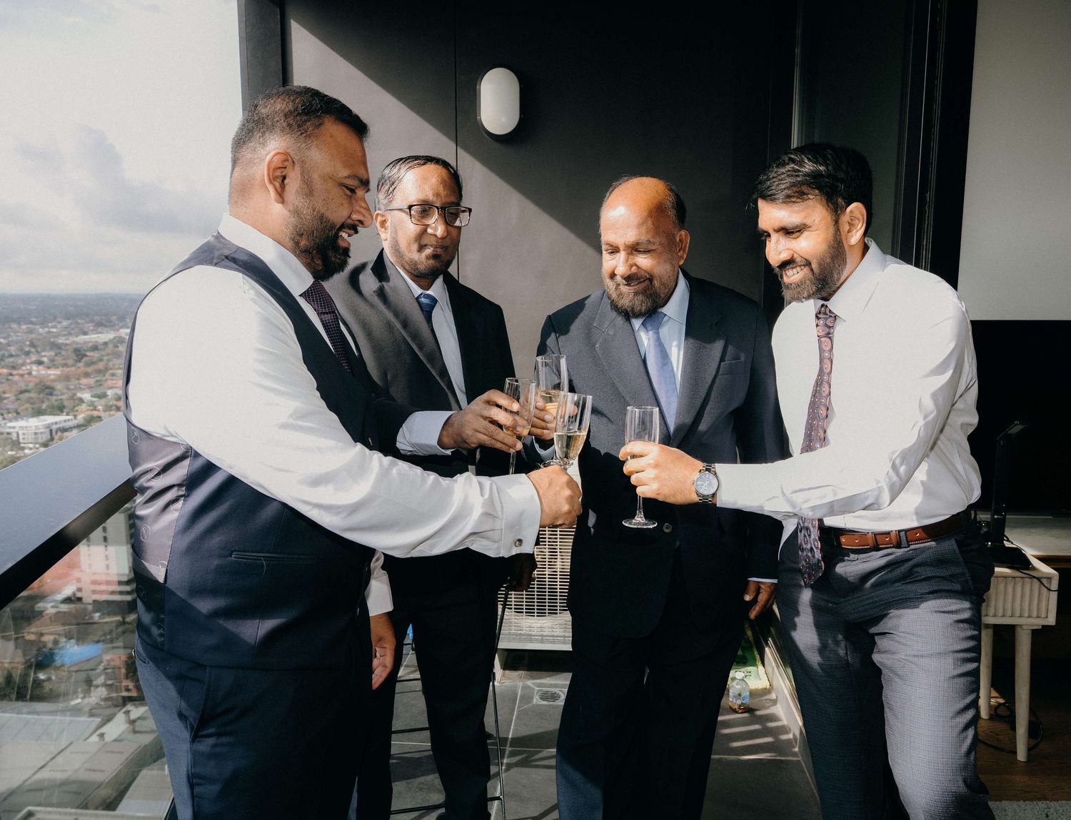 Four businessmen in formal attire toast with wine glasses on a balcony overlooking a cityscape view.