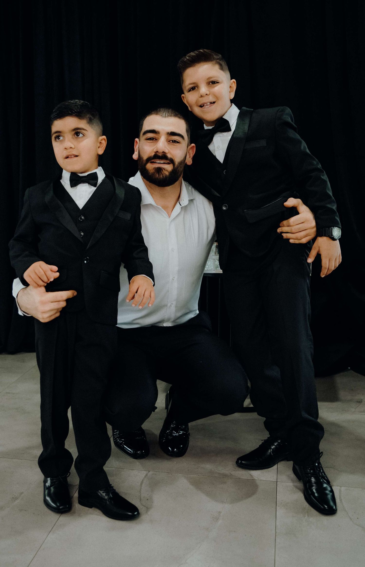 Three people in formal attire pose together, with matching black suits and bow ties on a light colored floor.