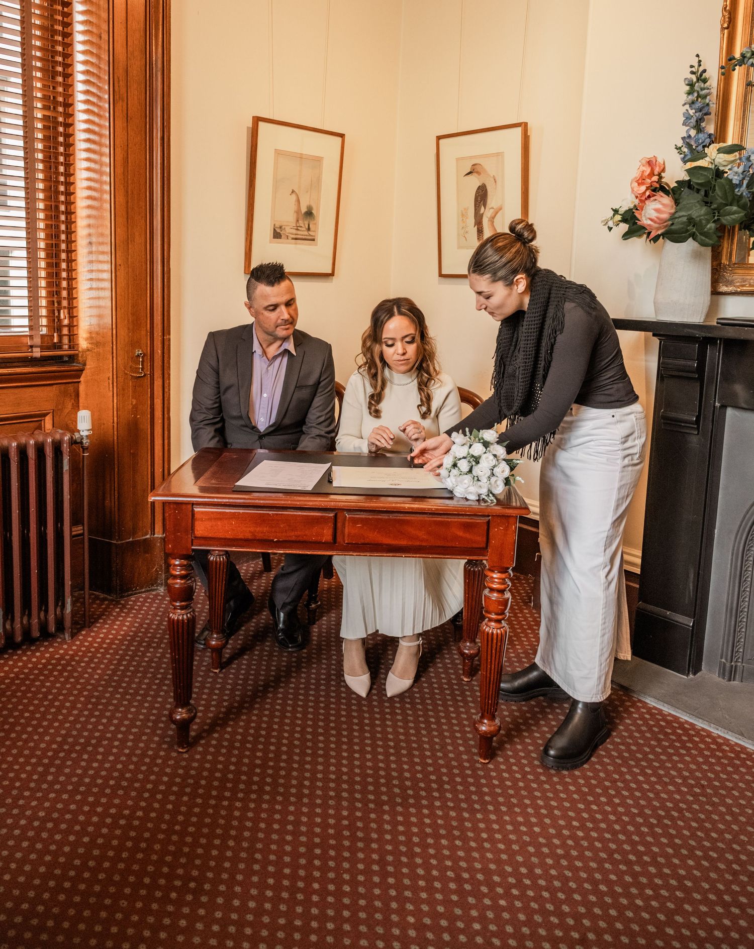 Three people gather around an antique wooden desk in a vintage-style room with patterned carpet and wooden blinds.
