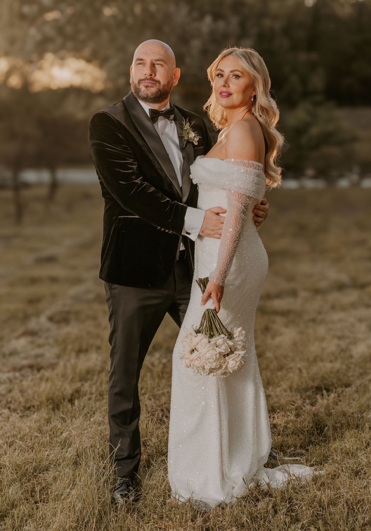 A couple shares an intimate moment in formal wedding attire while standing in a sunlit grassy field at golden hour.