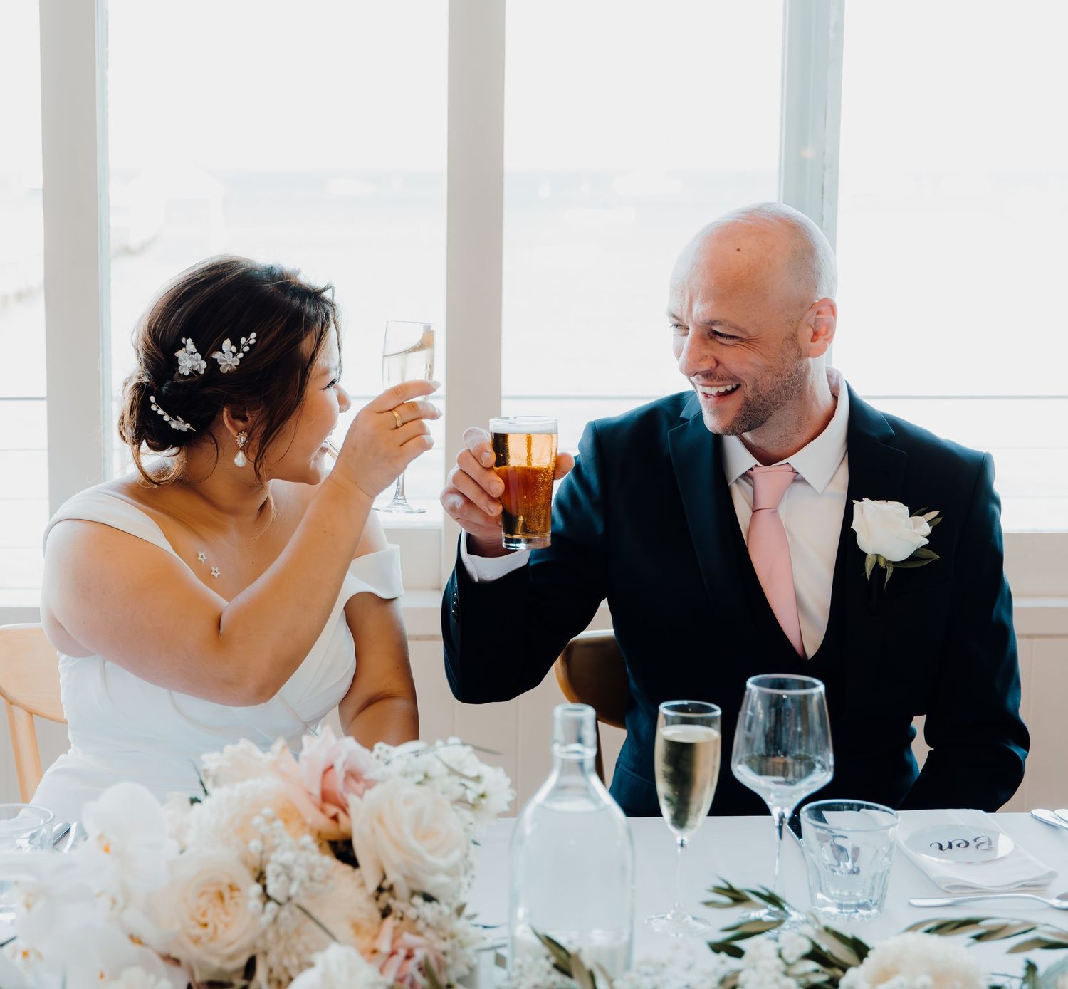 Wedding guests raise glasses in celebration during a bright reception dinner at a white-tablecloth venue.