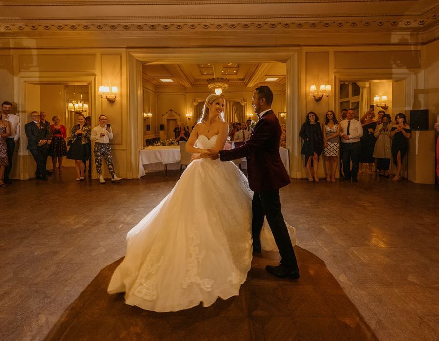 A couple dances in an elegant ballroom with ornate chandeliers and golden wall sconces during a wedding celebration.