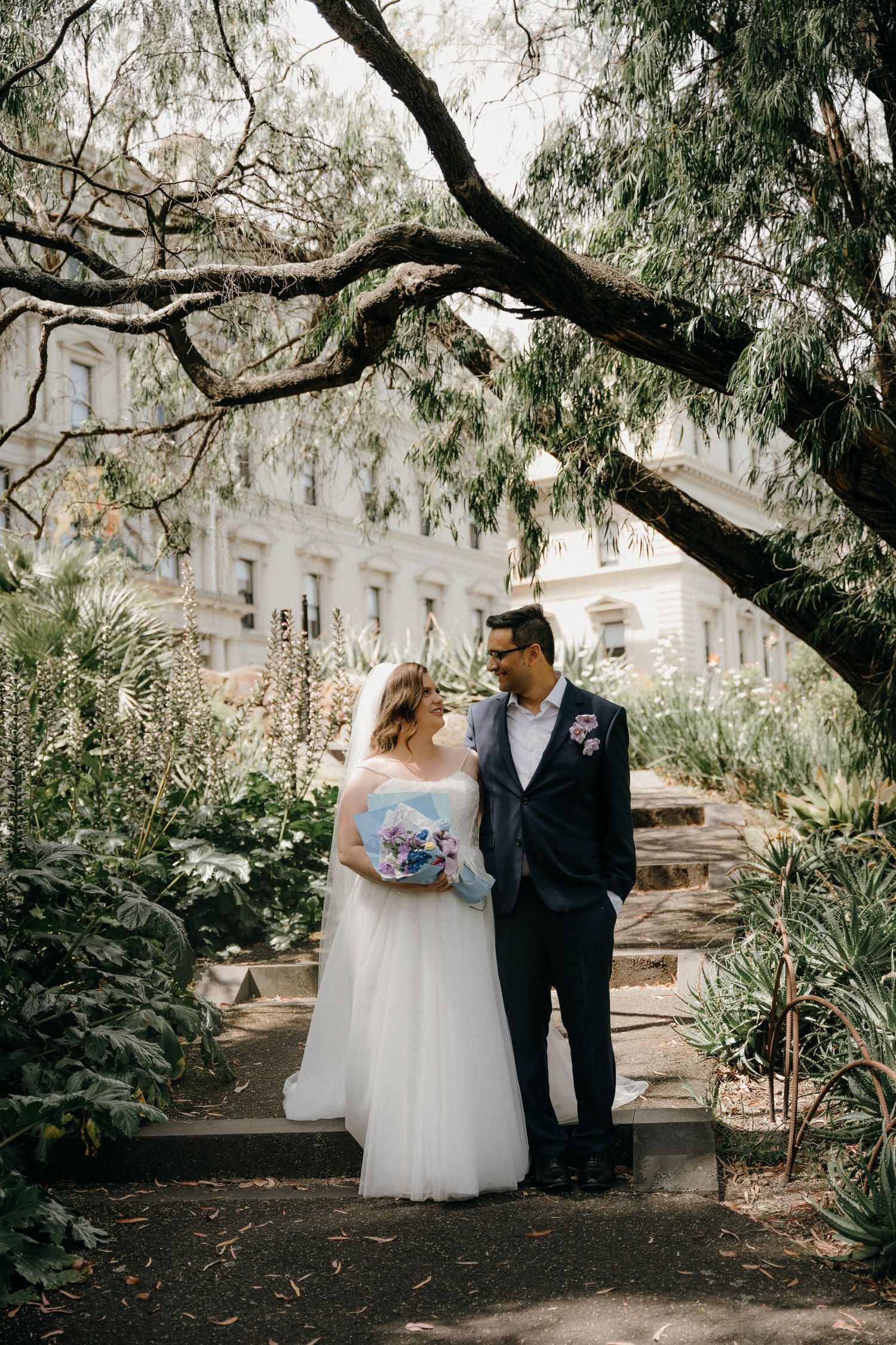A romantic wedding couple shares intimate moments under overhanging trees in a garden setting.