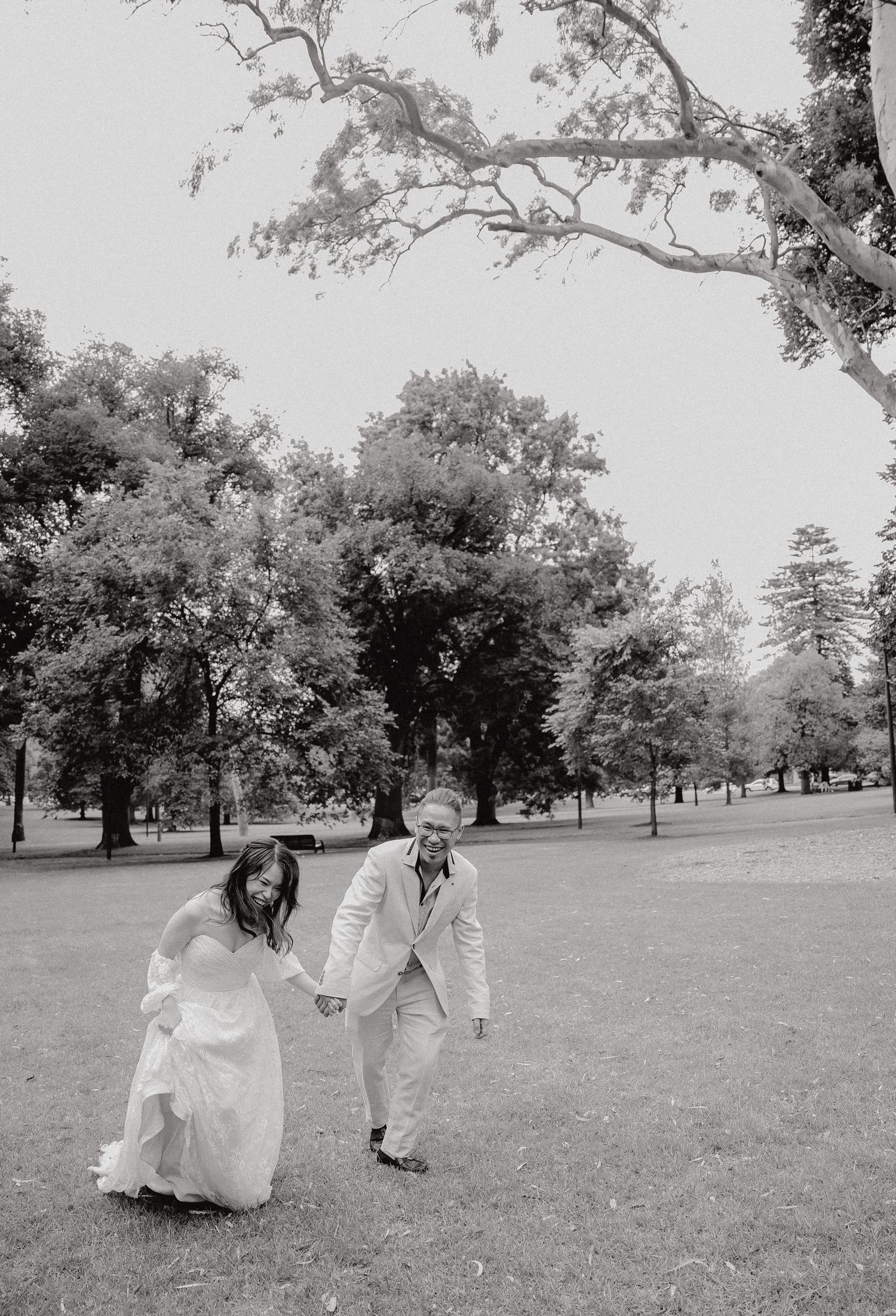 A couple in wedding attire walks and plays together on a grassy lawn in a park setting, captured in black and white.