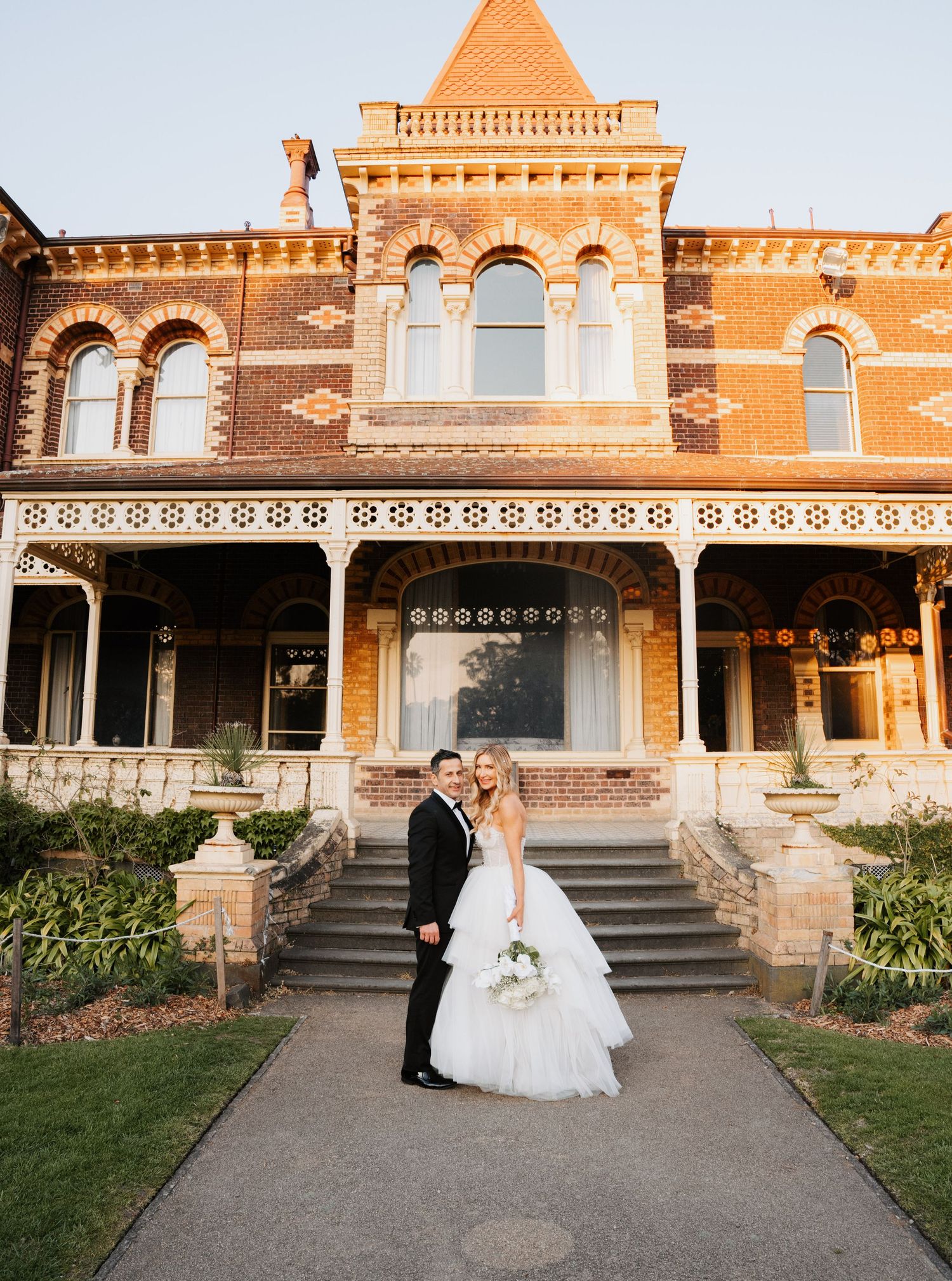 A couple stands in front of a historic Victorian mansion with ornate architecture and a grand staircase on their wedding day.