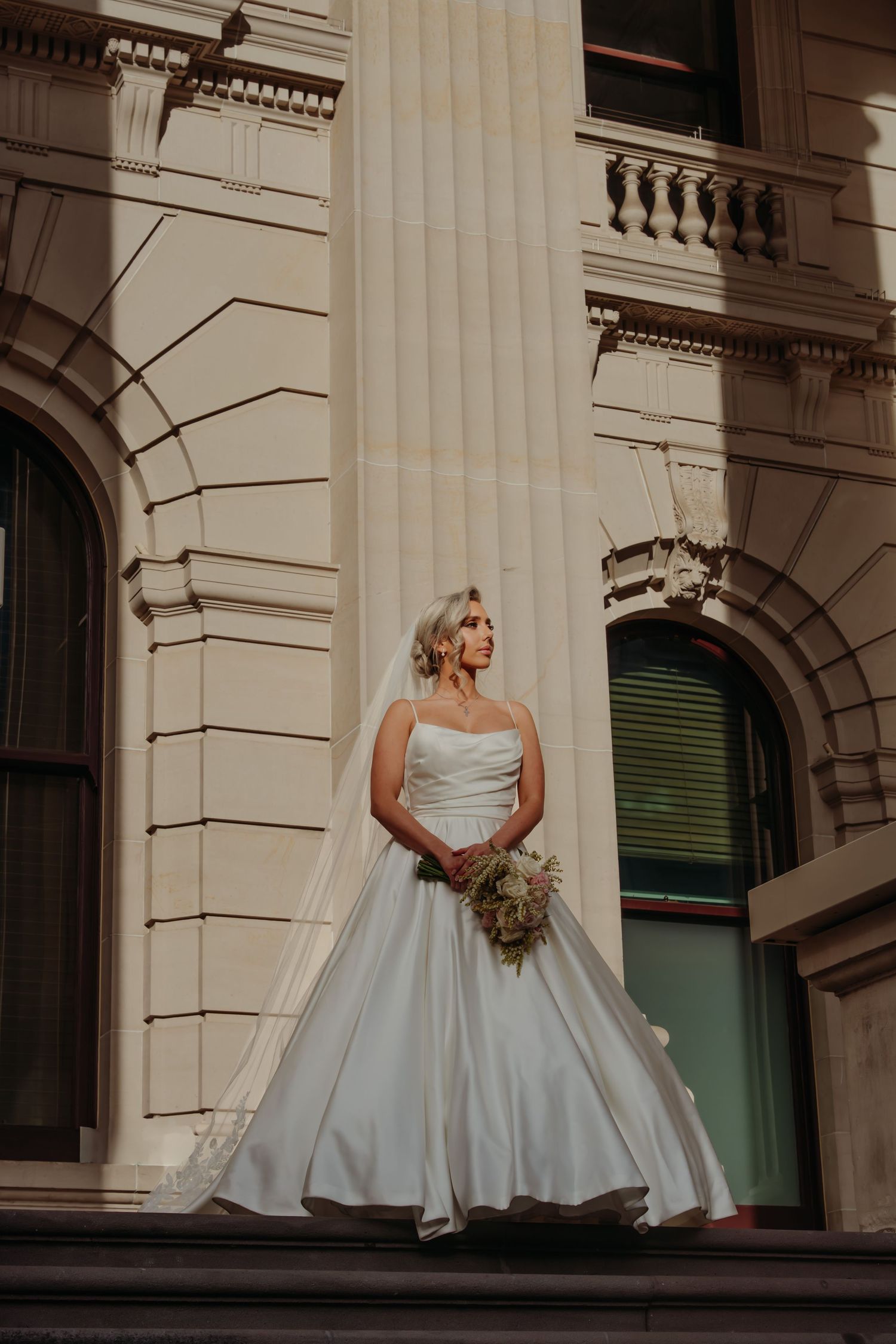A bride in a flowing white gown poses elegantly between classical stone columns of a grand building.