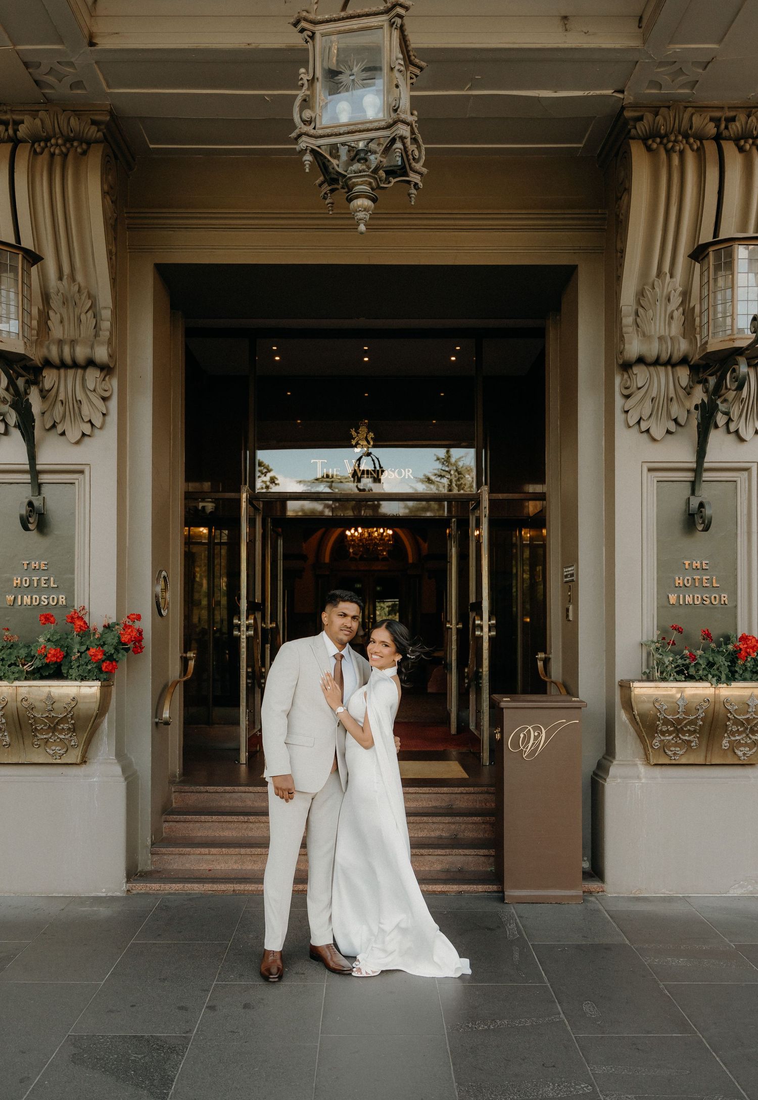Elegant wedding couple poses in front of Hotel Monaco entrance with white and beige decor and red flowers.