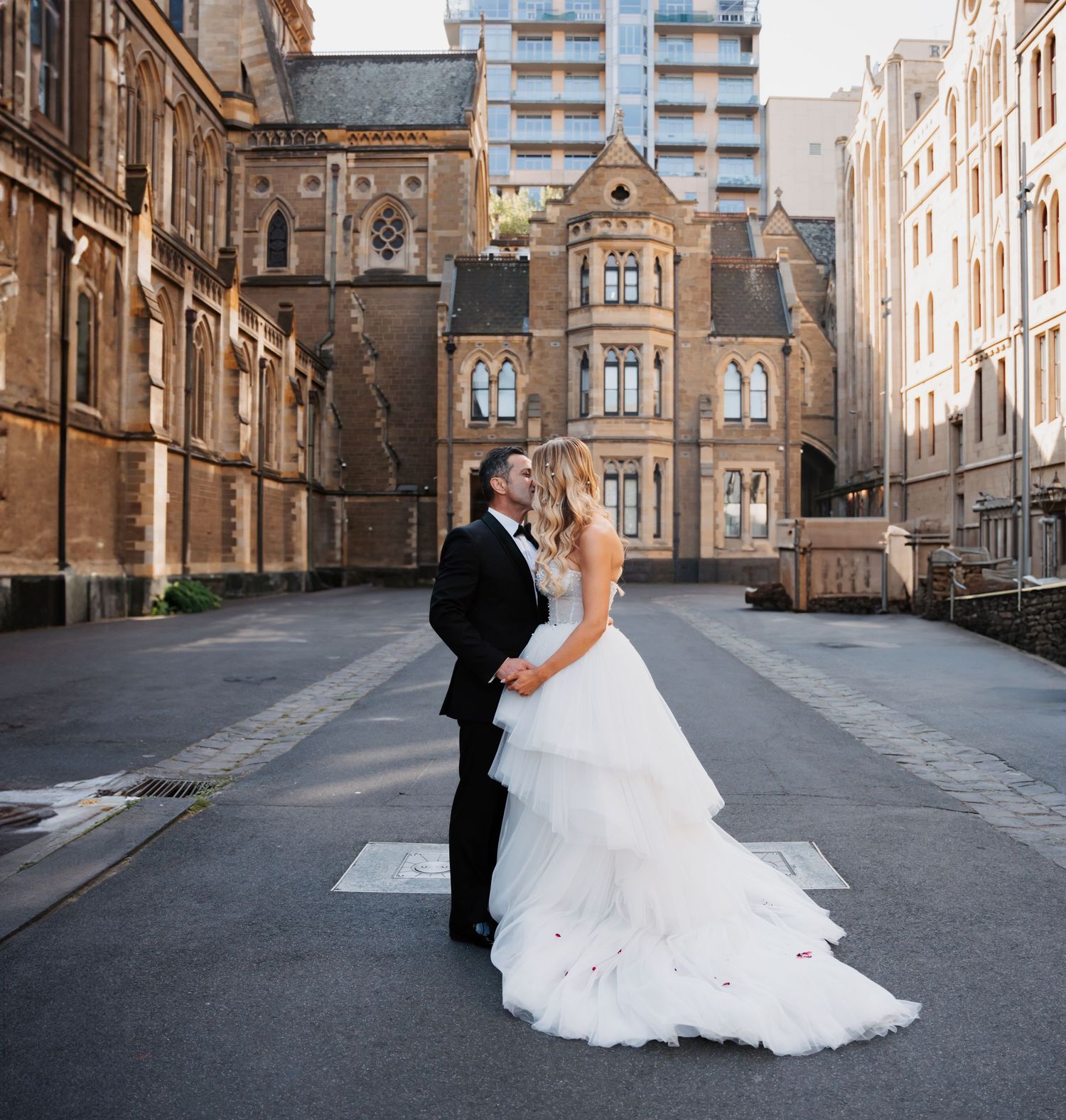 A couple in wedding attire share an intimate moment on a city street with historic Gothic architecture in the background.