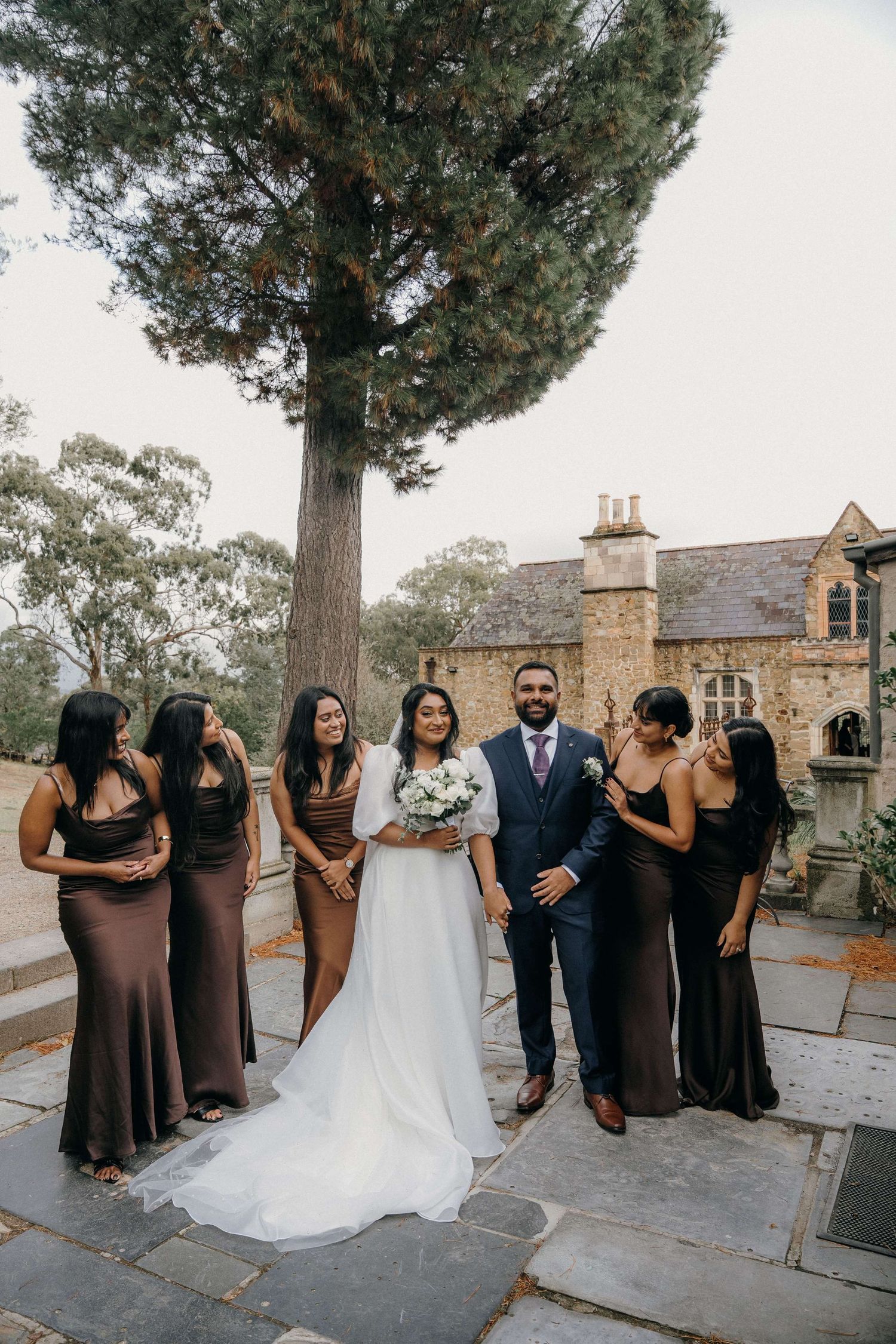 Wedding party stands in line outdoors on terrace with stone buildings and trees in background.