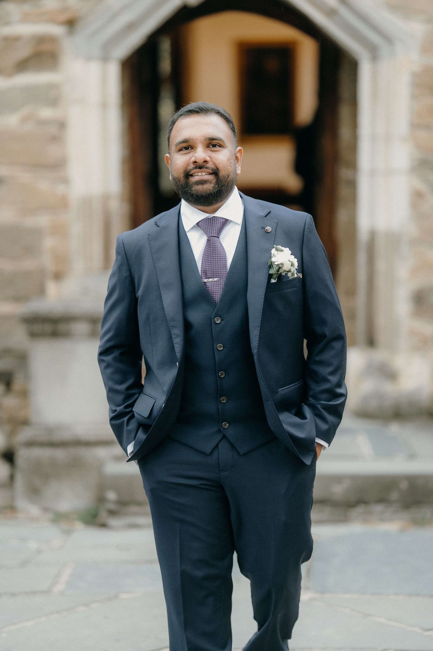 A groom in a stylish navy three-piece suit with purple tie and boutonniere smiles while standing outside a stone building.
