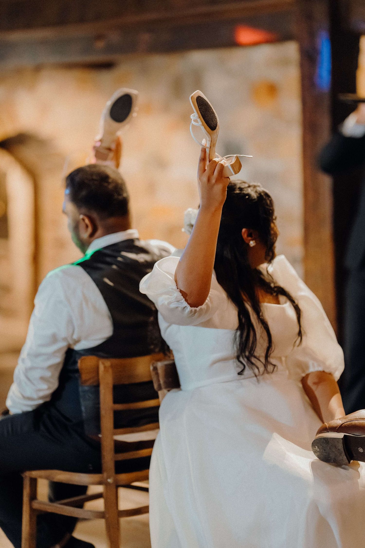 A couple at their wedding reception sit at a bar holding up phones while taking selfies in a warmly lit venue.